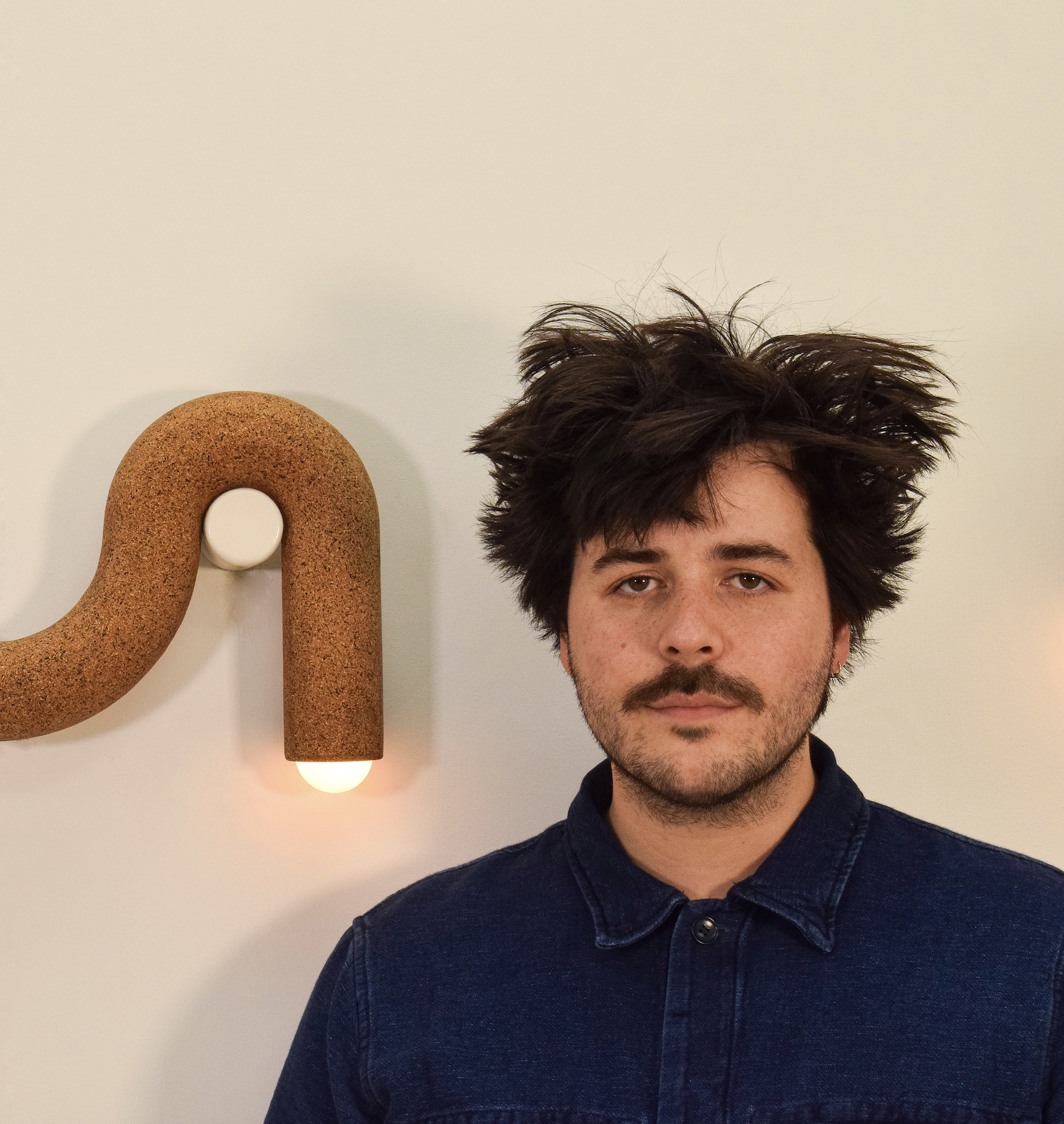 A man with dark, messy hair and facial hair wearing a dark denim jacket, standing in front of a white wall with a textured, brown, bowtie-shaped light or sculpture and a circular white element beside it.