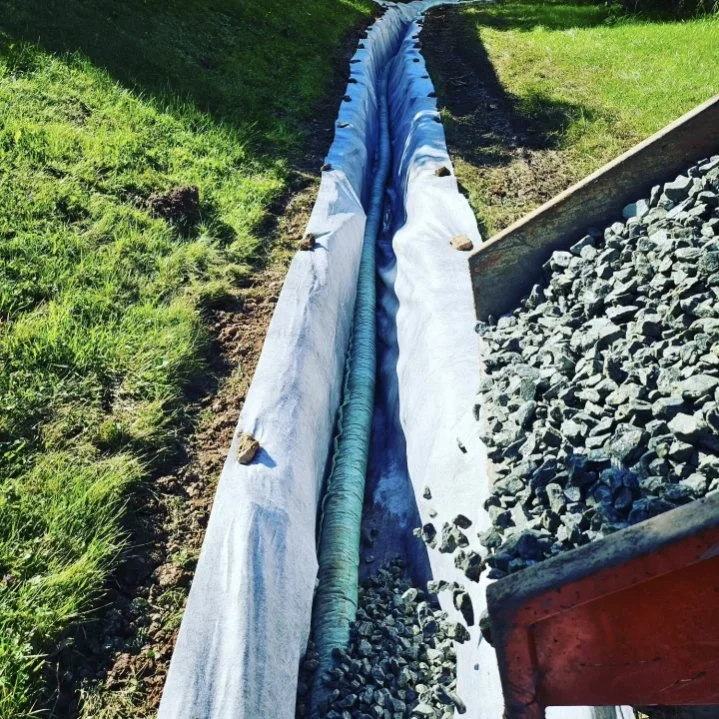 Construction site showing gravel being poured into a fabric-lined trench, possibly for drainage installation, surrounded by grass.