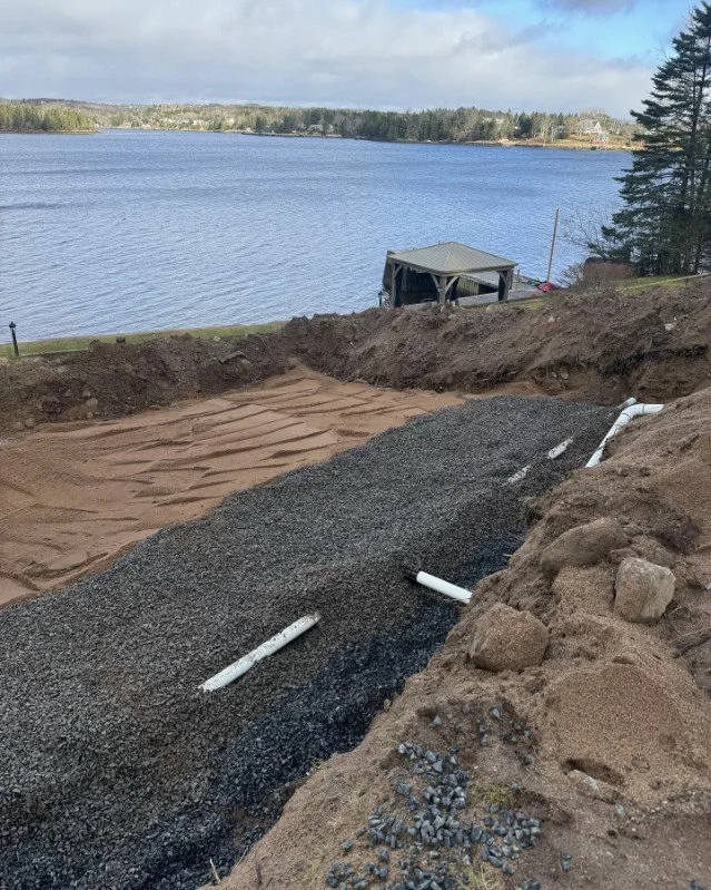 Excavated area with gravel and pipes near a lake with a small dock and trees in the background.