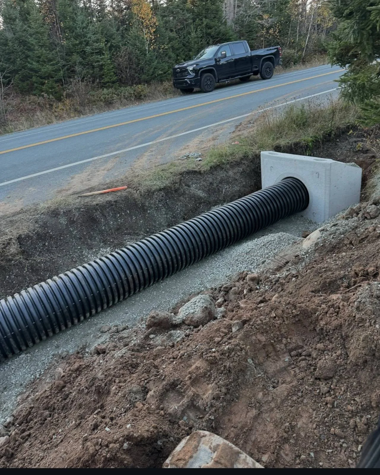 A black pickup truck parked on the road near a construction site with a large drainage pipe embedded in the ground, surrounded by dirt and gravel, and bordered by trees.