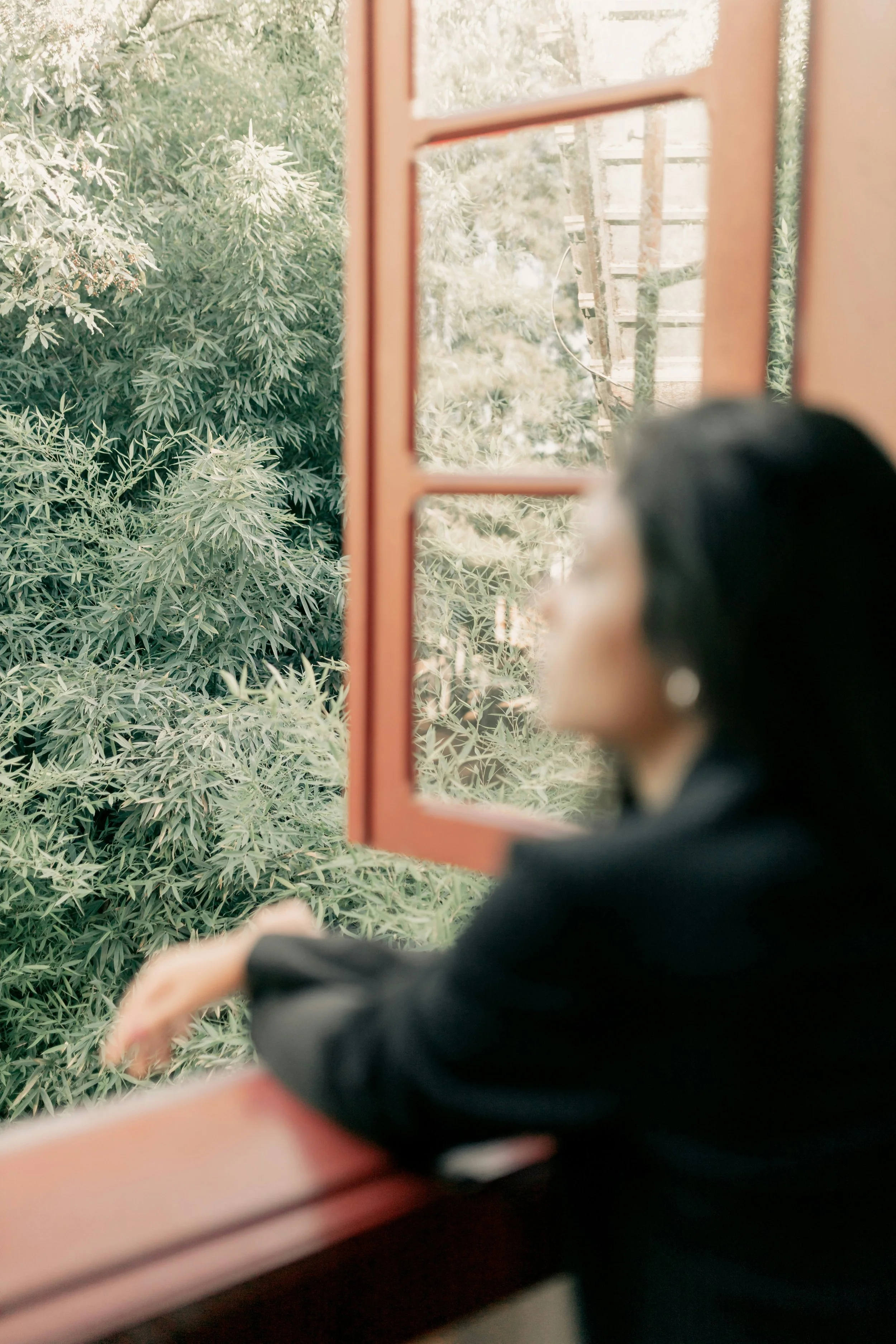 A woman looking out of a reddish-orange window at lush green trees outside.