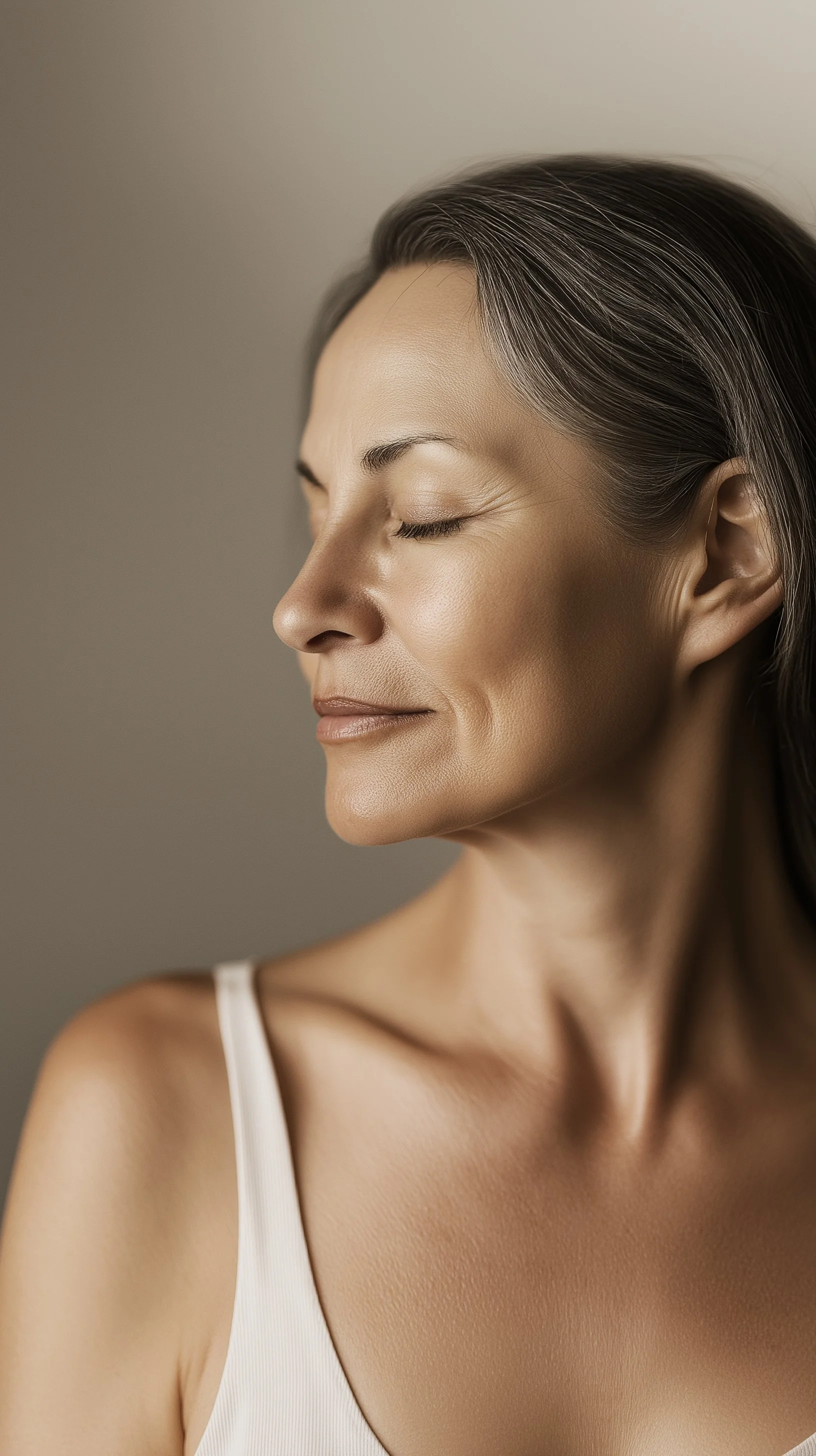 A mature woman with gray hair and smooth skin, eyes closed, wearing a white tank top and smiling peacefully.