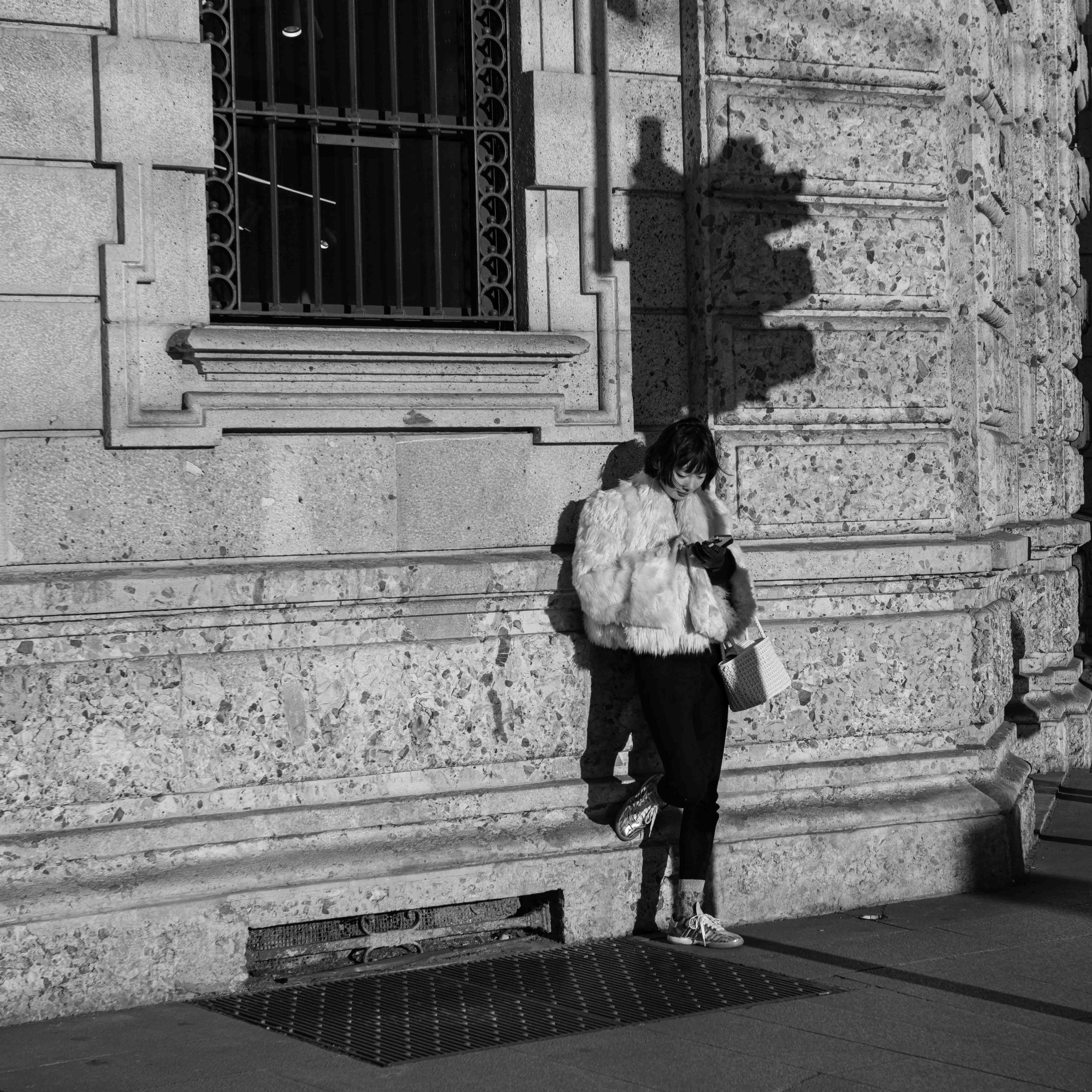 The world moved, but she stayed still.
She leaned effortlessly against the stone wall, her fur coat catching the light, her metallic boots glinting with each shift of her weight. The sun sculpted her silhouette, the building’s ornate details framing