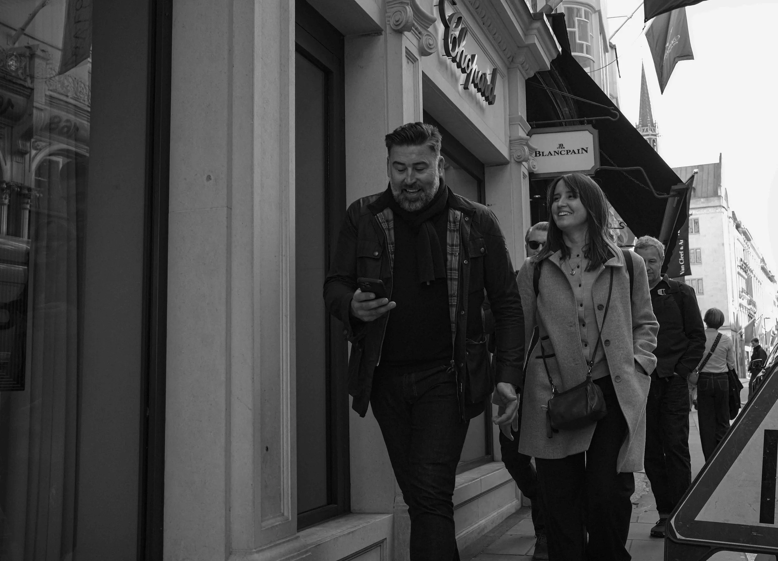 Laughter spills into the street as he reads something amusing on his phone. She listens, her eyes reflecting curiosity and delight. They walk in sync, their casual elegance blending into the sophisticated cityscape.
Behind them, life continues—stran