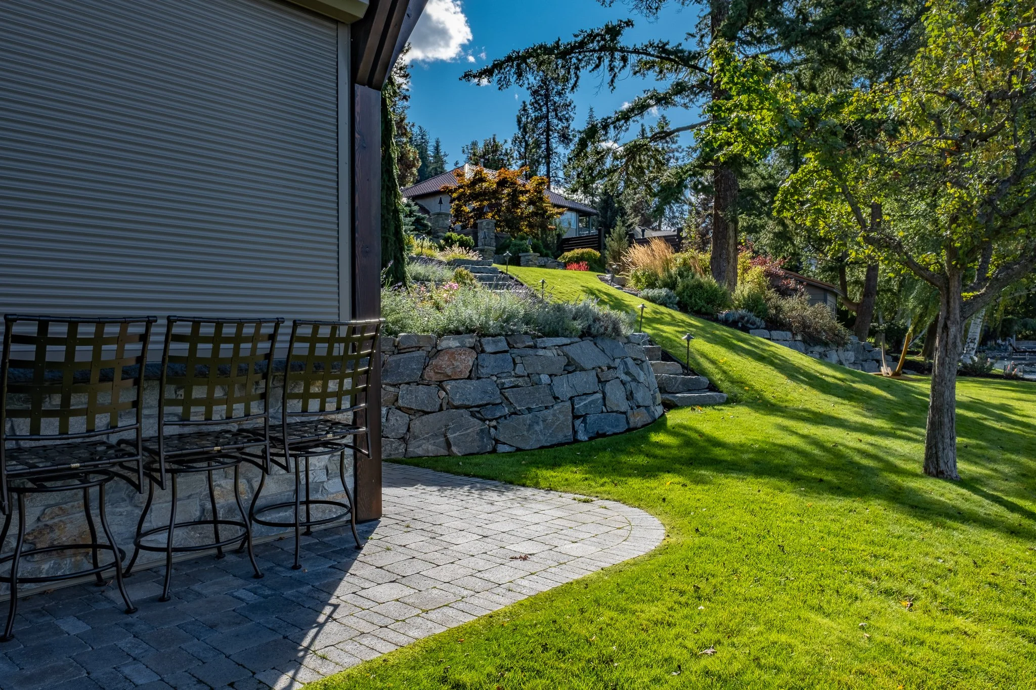 Backyard scene with stone bar chairs, stone retaining wall, green lawn, trees, and a house in the background.
