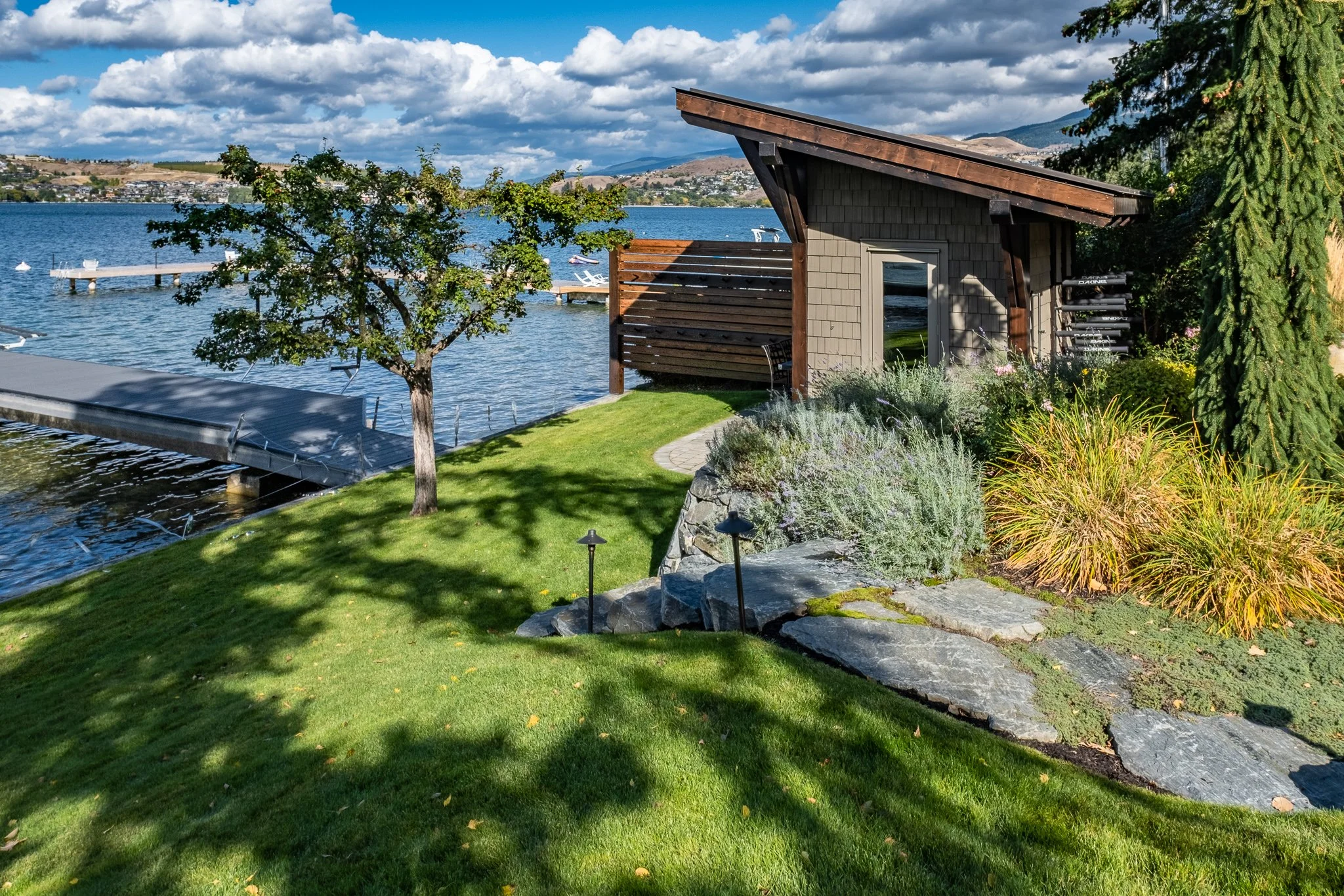 Lakeside house with a sloped lawn, a wooden dock extending over the water, trees, and landscaped garden, under a blue sky with clouds.