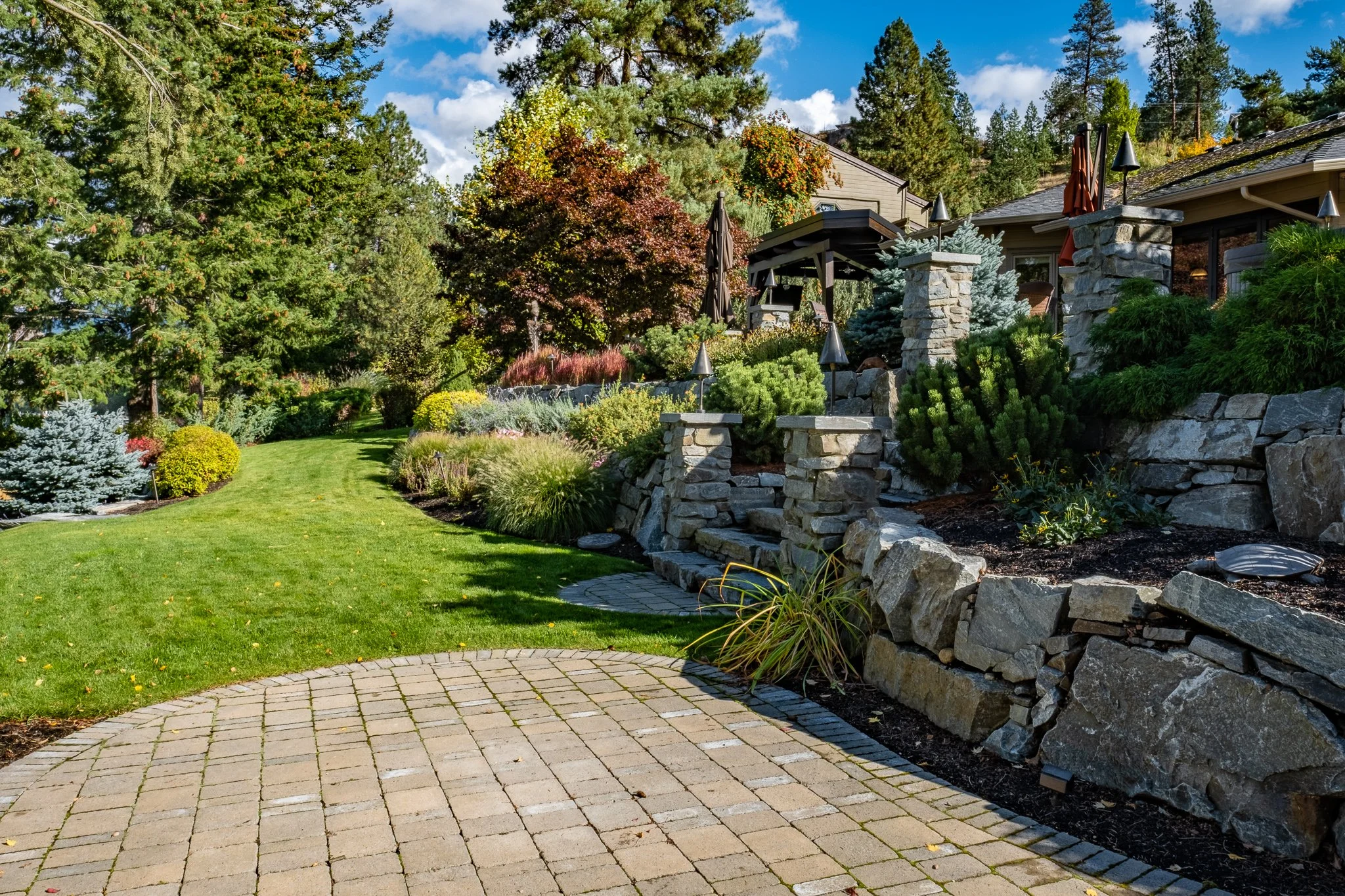 Landscaped garden with stone pathway, green lawn, shrubs, and trees next to a house.