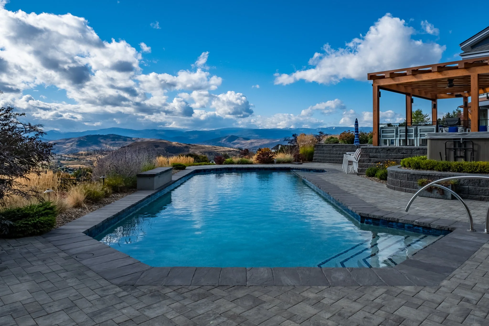 A rectangular outdoor swimming pool with a paved deck, overlooking scenic hills and mountains under a cloudy sky. A wooden pergola is on the right, adjacent to patio furniture.