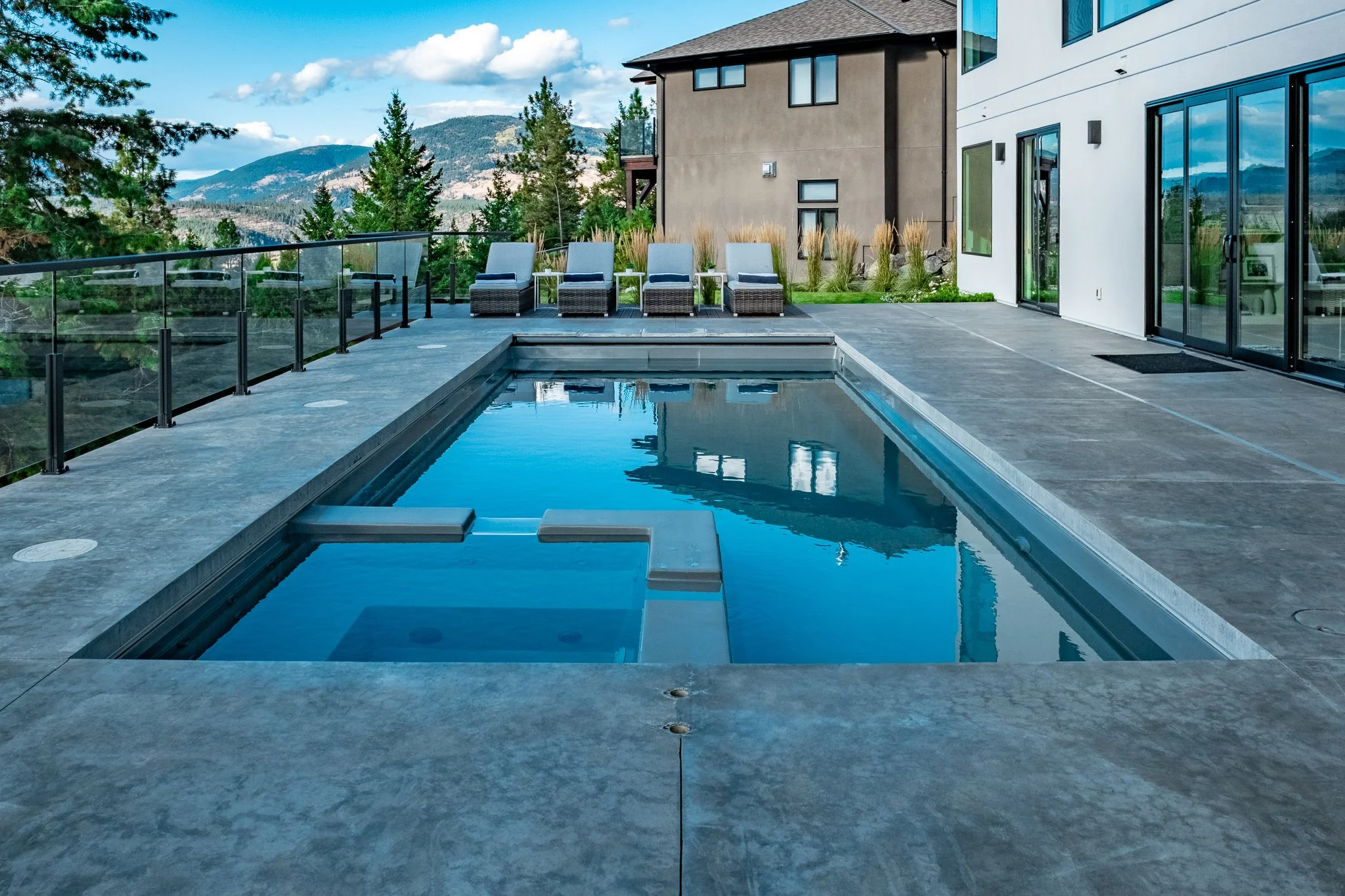A modern outdoor swimming pool with lounge chairs on a concrete deck, surrounded by glass railings, near a contemporary house and trees.
