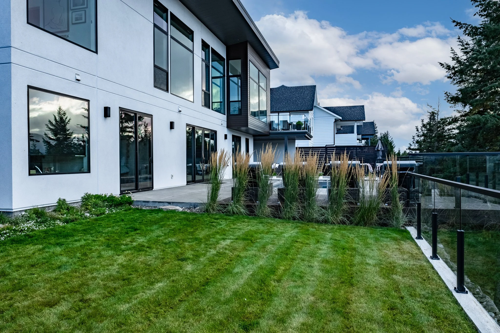 Modern house exterior with large windows, patio, and landscaped lawn with ornamental grasses.