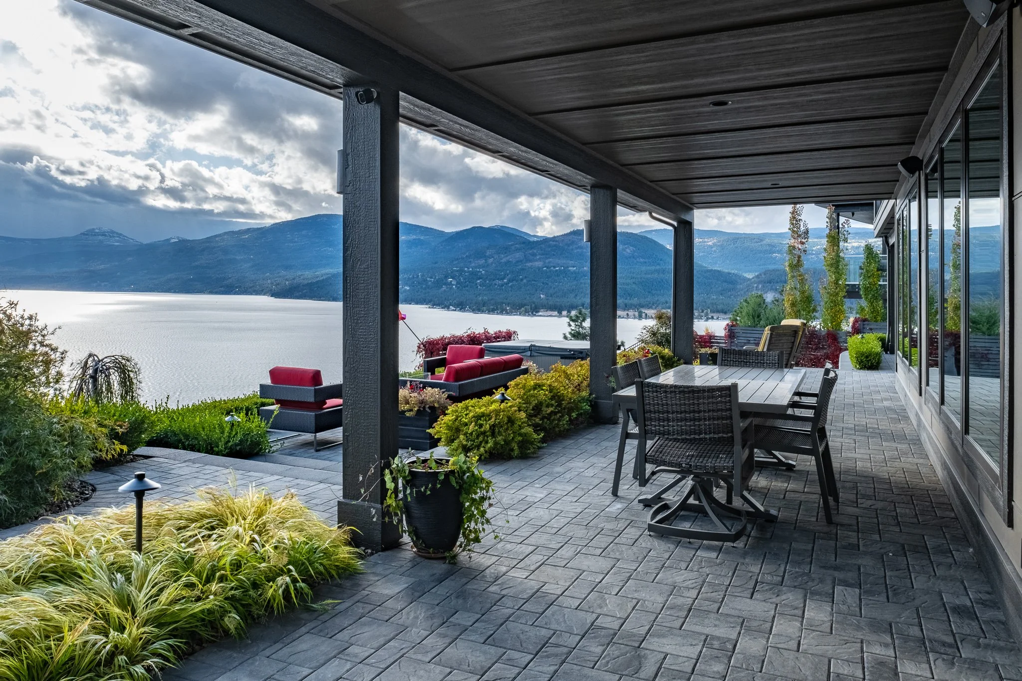 Covered patio with outdoor furniture, overlooking a lake and mountains, surrounded by lush greenery and plants.