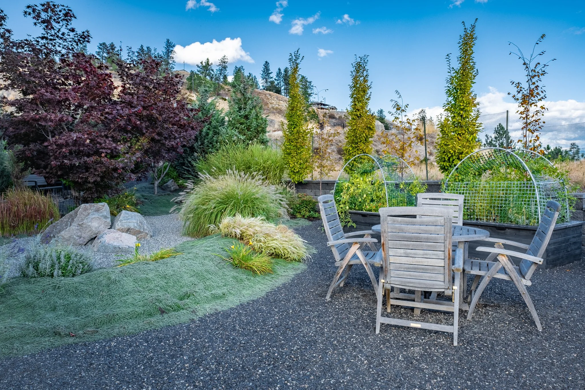 Outdoor garden with a round wooden table and chairs, surrounded by lush greenery, trees, and decorative rocks under a clear blue sky.