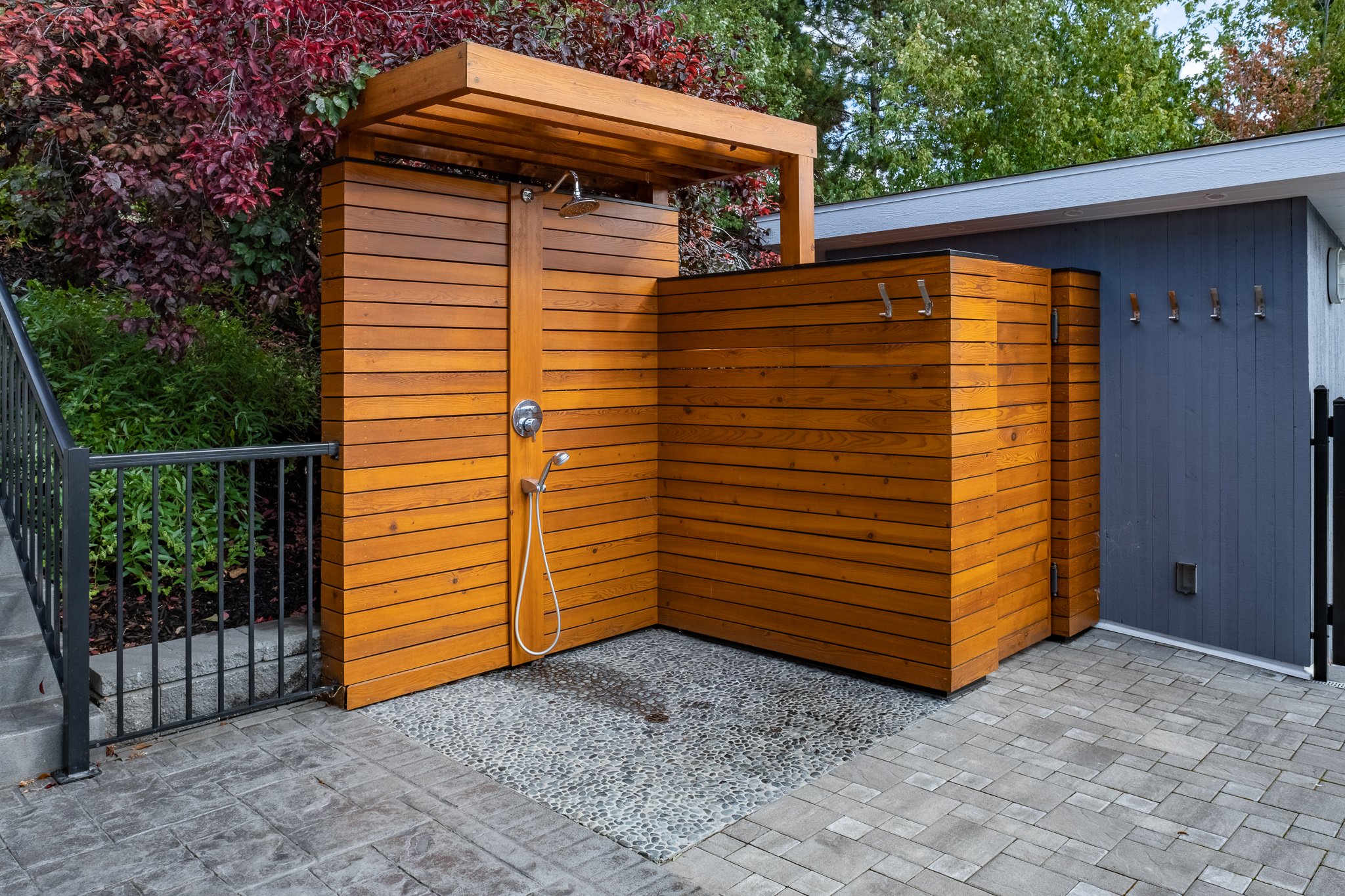 Outdoor wooden shower with privacy screen, paved brick floor, and surrounding greenery.
