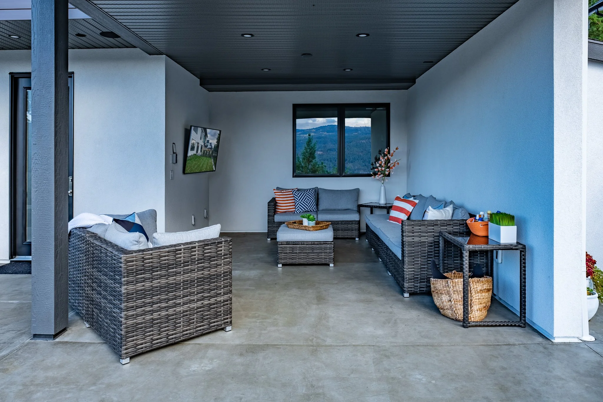 Modern outdoor patio area with gray wicker furniture, blue cushions, striped pillows, a wall-mounted TV, and a view of mountains through a window.