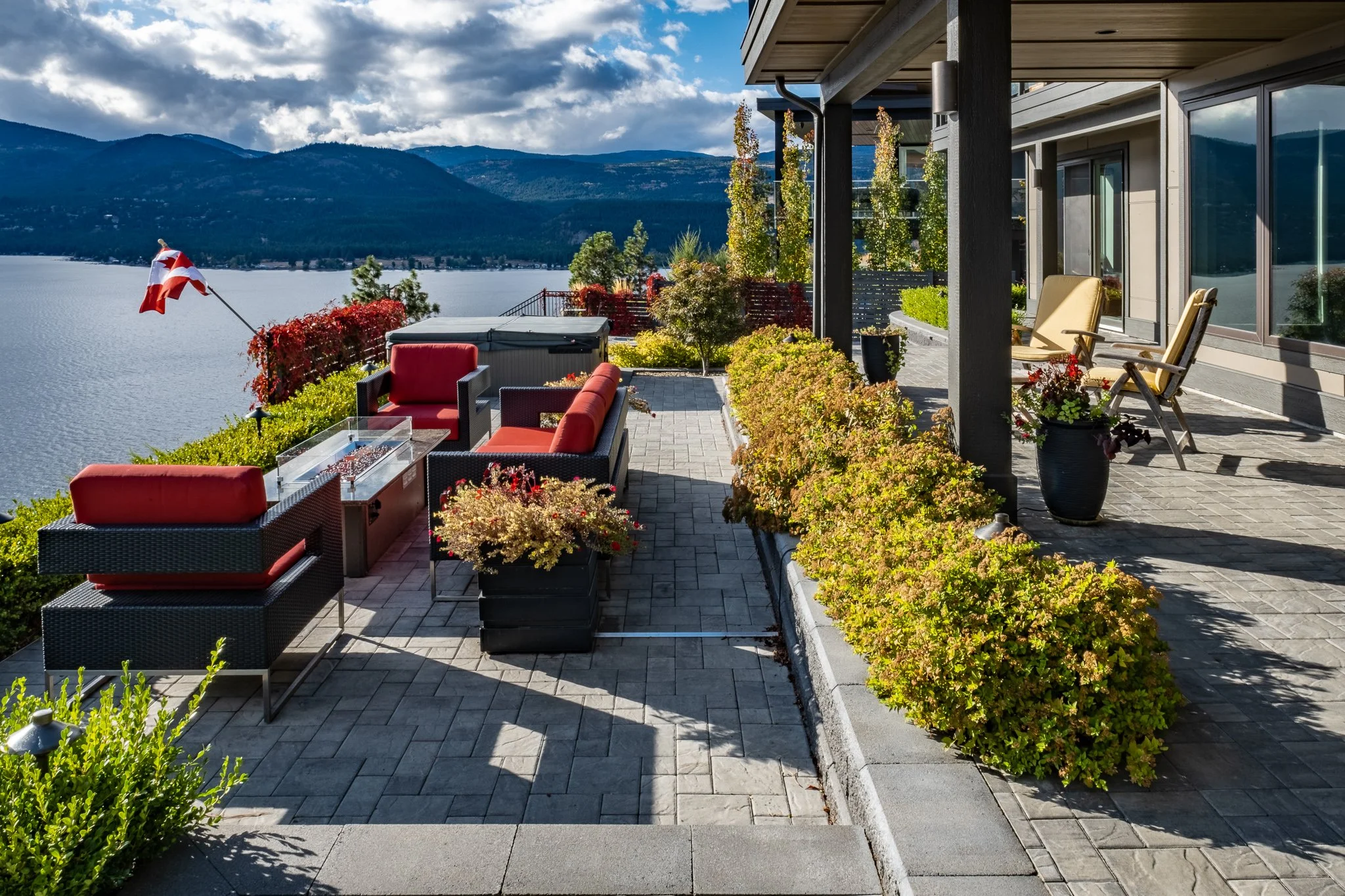 Lakeside patio with red cushioned seating, fire pit table, Canadian flag, and panoramic mountain and water views.