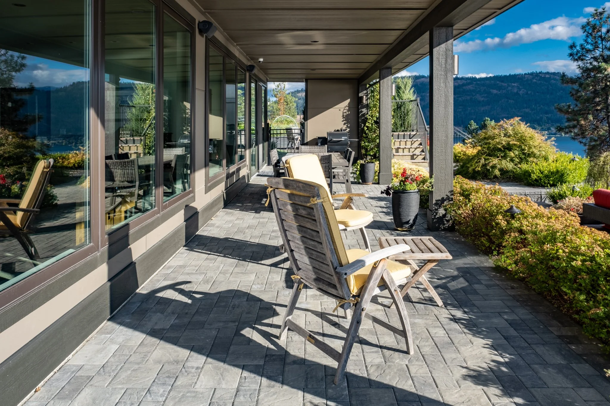 Patio area with wooden chairs, small tables, colorful potted plants, overlooking a scenic landscape with hills and water.