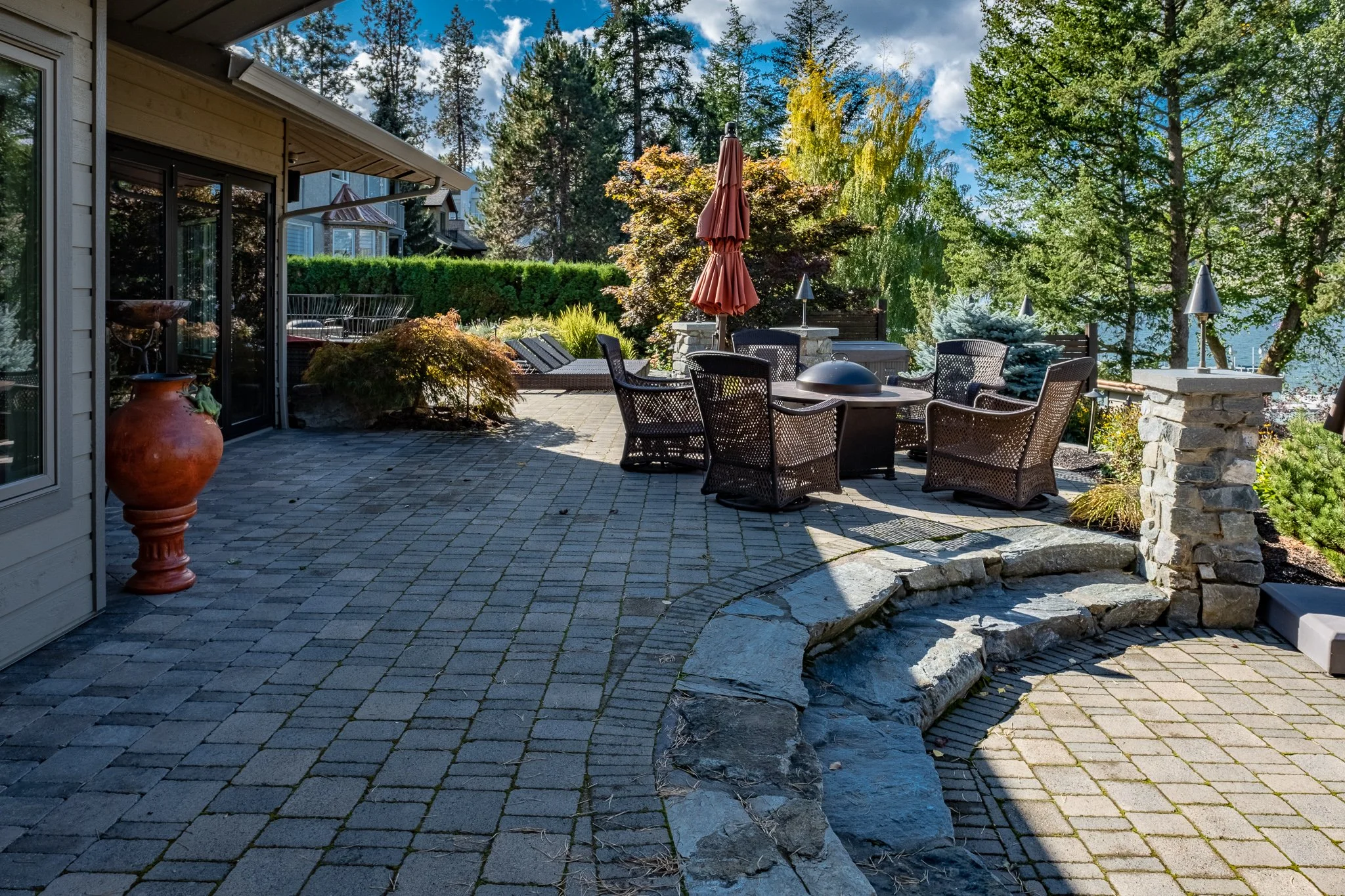 Outdoor patio with wicker furniture, a round table, and a closed umbrella. The area is surrounded by trees and plants, with stone tile flooring. A stone pathway and large pot are also visible.