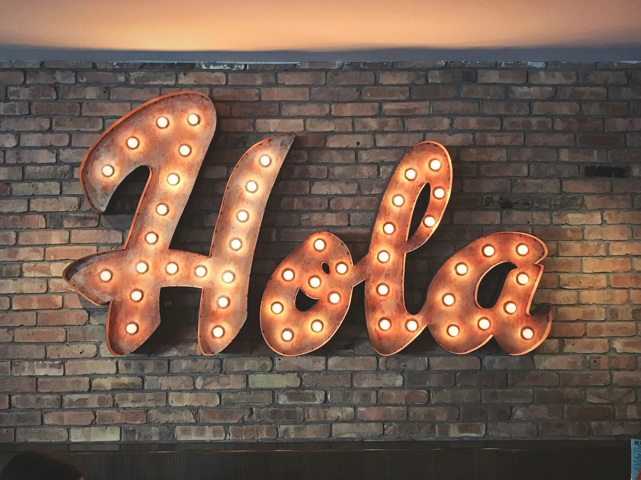 Decorative large marquee sign spelling out 'Hola' with light bulbs, mounted on an exposed brick wall inside a restaurant or café.