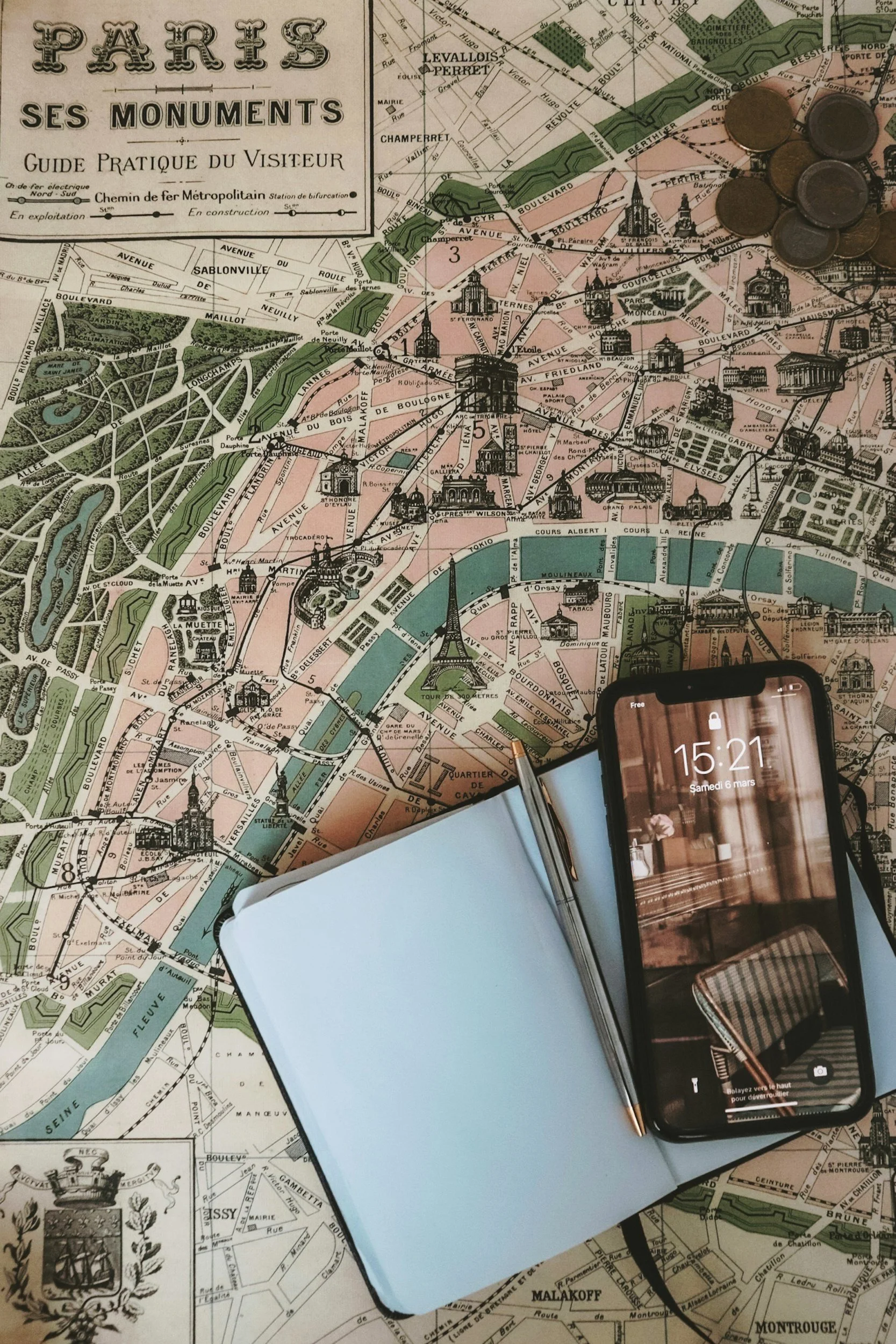 A detailed map of Paris with an open notebook, a pen, a smartphone displaying a photo of an interior, and several coins placed on the map.