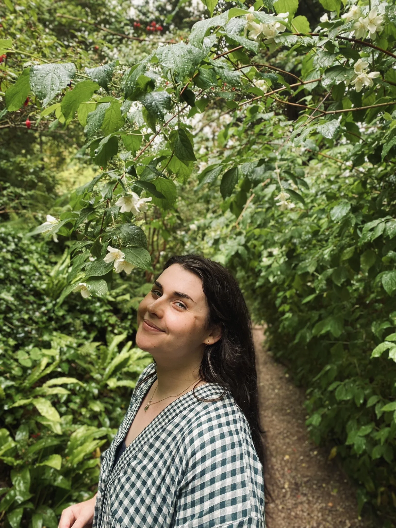 A woman with dark hair and a checked shirt smiling outdoors in a lush, green garden with flowering plants and a narrow dirt path.