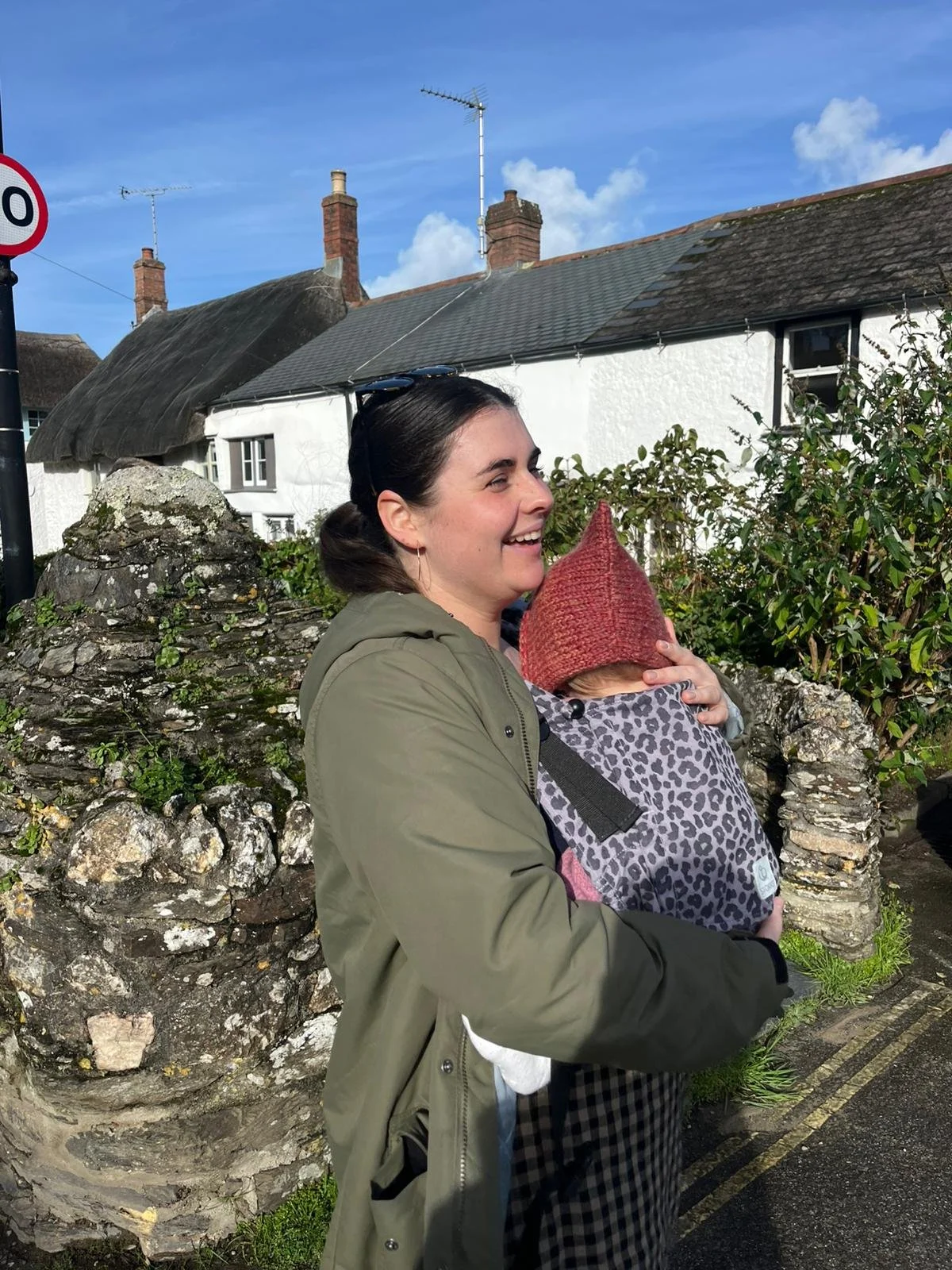 A woman with dark hair in a ponytail wearing a green jacket hugging a child in a red knit hat and leopard print coat outdoors in front of a stone wall, with houses and a blue sky in the background.