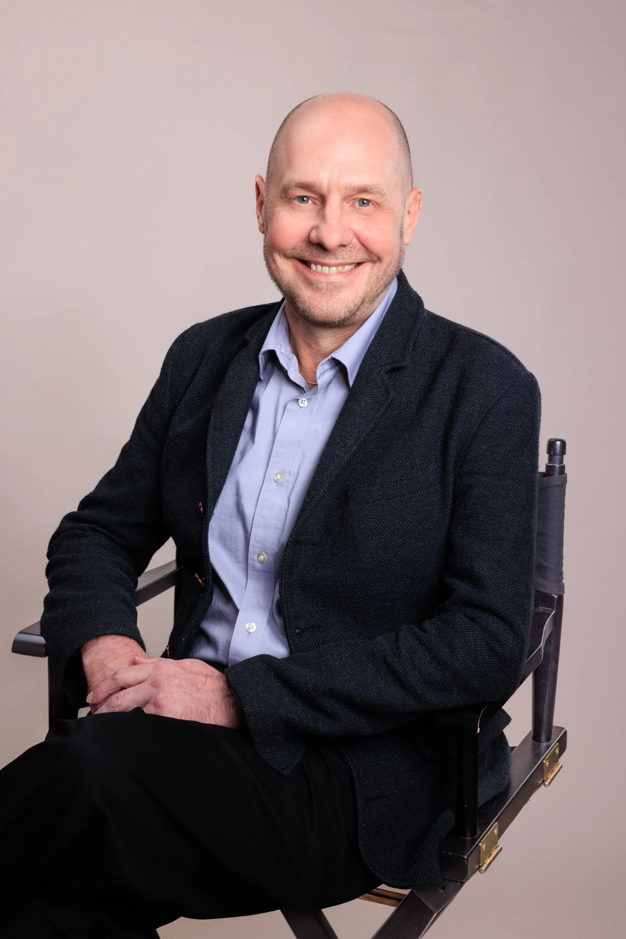 A smiling middle-aged man sitting on a director's chair, wearing a dark blazer, light blue shirt, and black pants, against a neutral background.