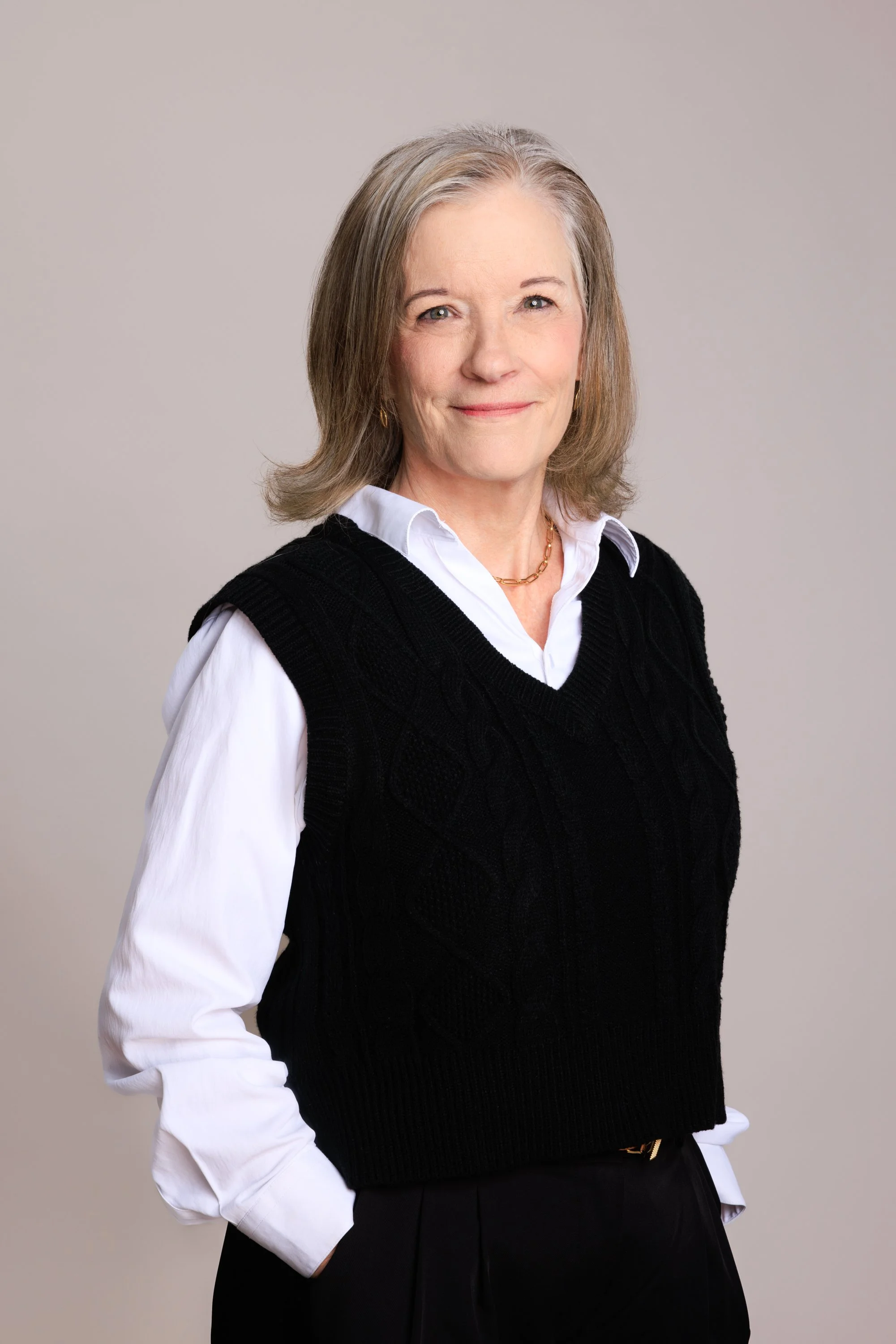 A portrait of a mature woman with gray hair, wearing a white collared shirt under a black sweater vest, with gold jewelry, standing against a plain light background.