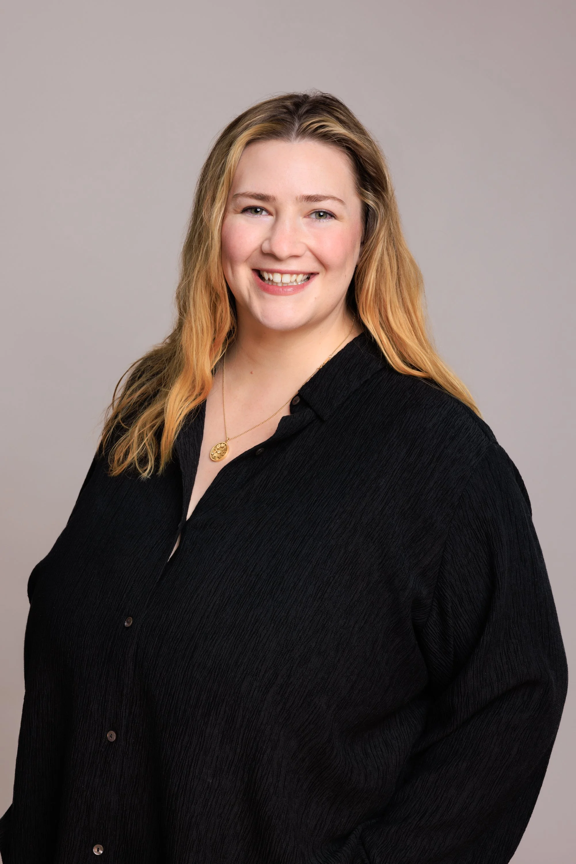 A woman with long wavy blonde hair wearing a black shirt and a gold necklace, smiling at the camera against a neutral gray background.