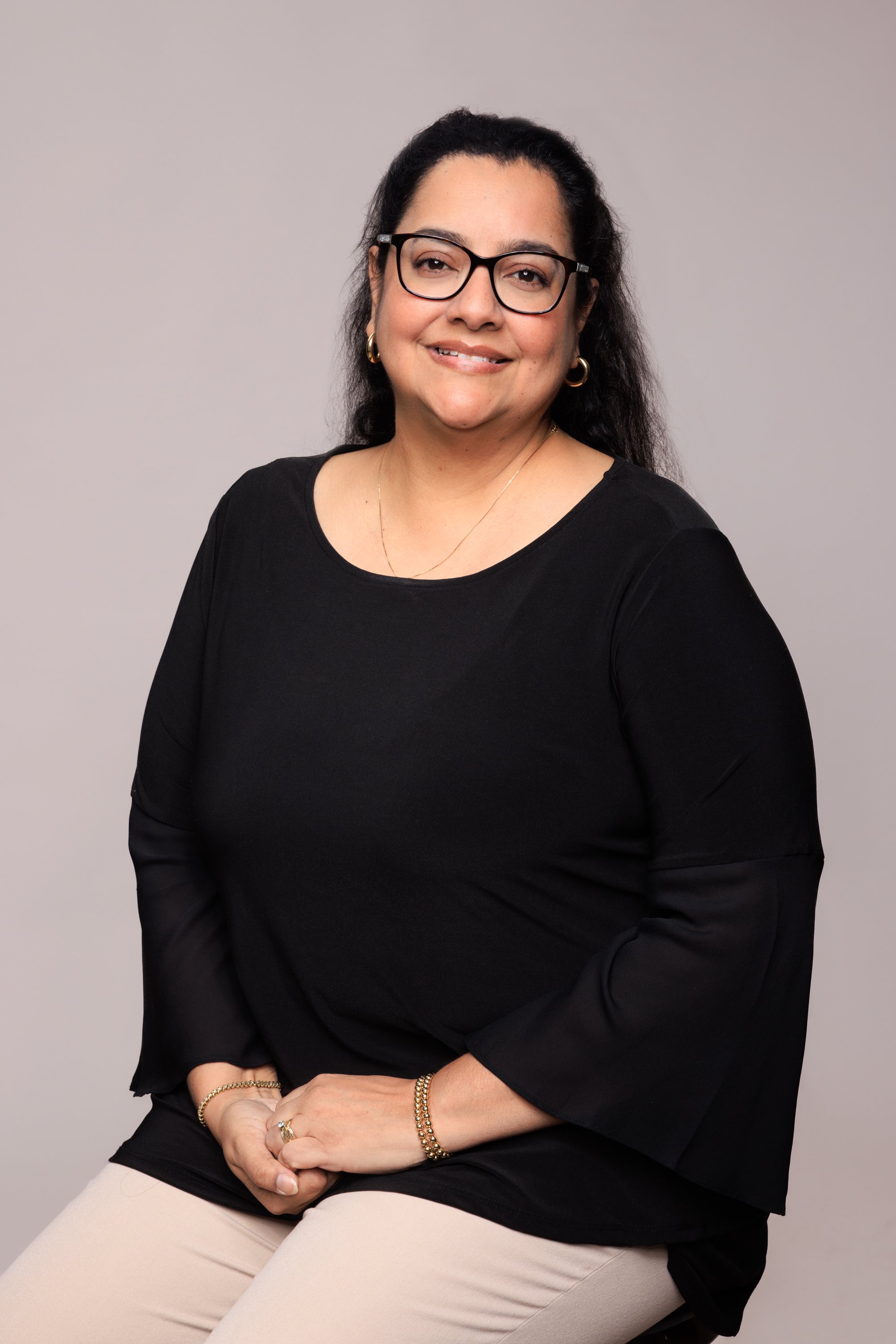 A middle-aged woman with dark hair, glasses, and jewelry, smiling and sitting against a plain gray background.