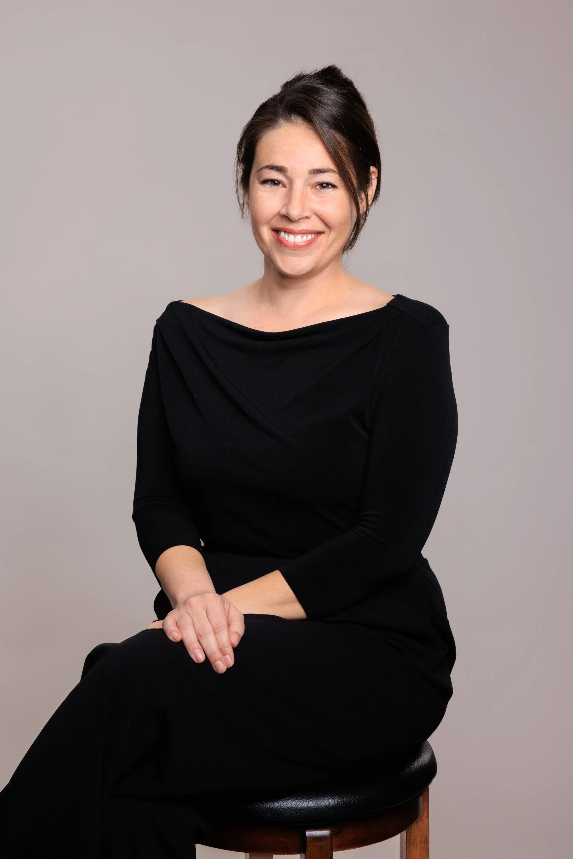 A woman with brown hair styled in an updo, wearing a black long-sleeve dress, sitting on a stool against a plain gray background, smiling and looking at the camera.