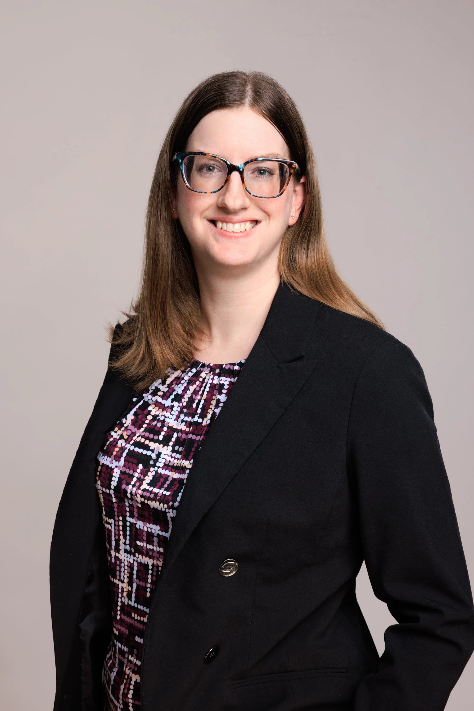 A professional headshot of a woman with shoulder-length brown hair, wearing glasses, a black blazer, and a patterned blouse, smiling against a plain neutral background.