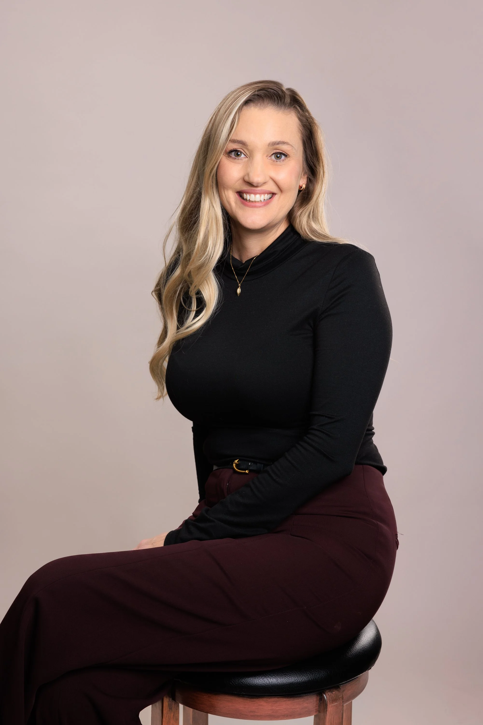 A woman with long blonde hair, wearing a black top and maroon pants, sitting on a stool with a neutral background, smiling at the camera.