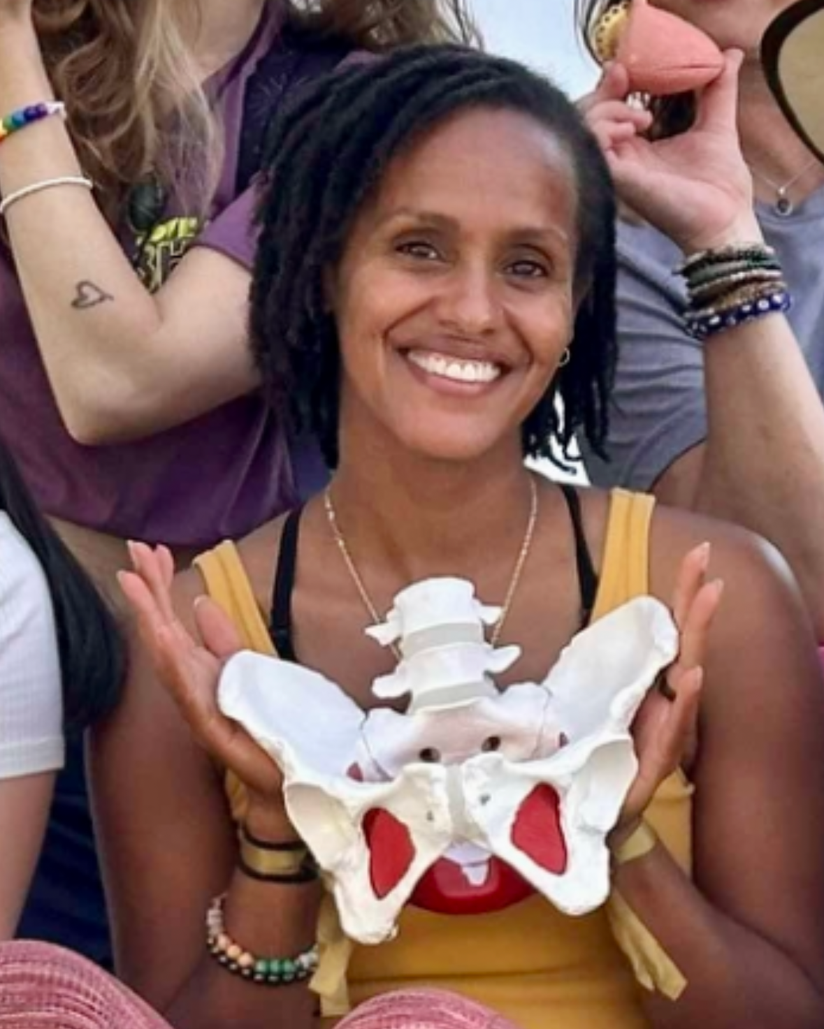 A woman with dreadlocks smiling and holding a model of a human pelvis and spine in front of her chest.