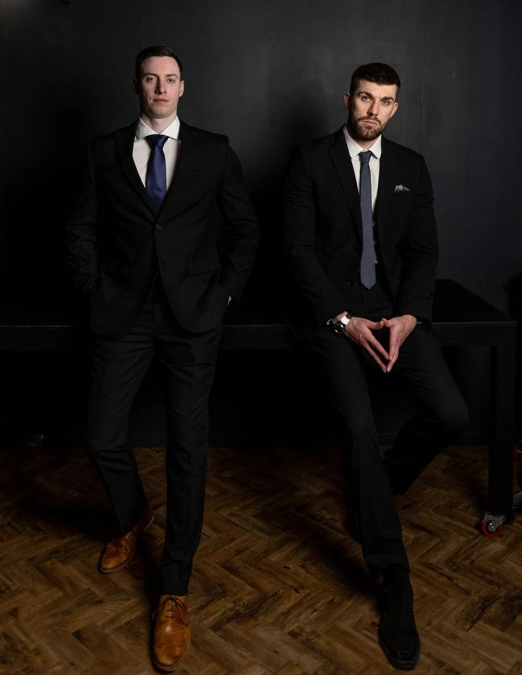 Two men in suits posing against a dark background, one standing with hands in pockets, the other sitting on a table.