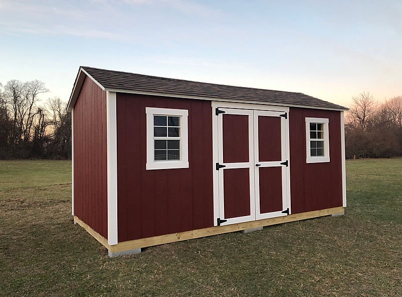 Red storage shed with white trim and two windows, positioned on a grassy field with trees in the background.
