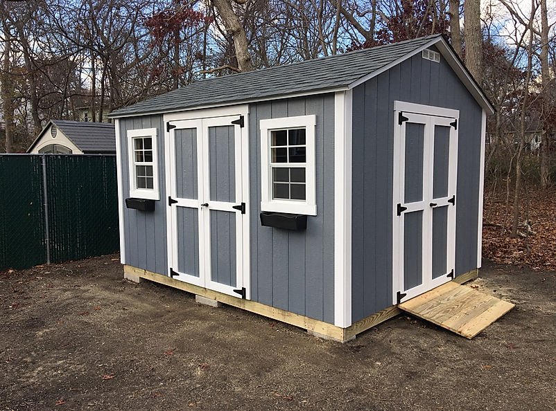 A small blue outdoor shed with white trim and two windows, situated in a wooded area.