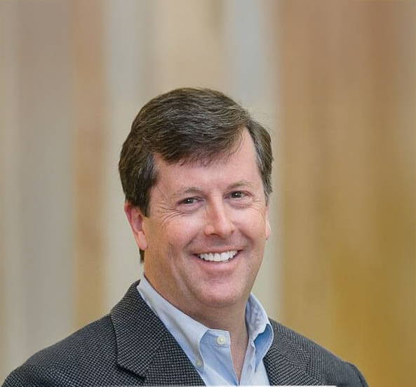 A man with brown hair smiling, wearing a dark blazer and light blue shirt, standing indoors with blurred beige curtains or walls in the background.