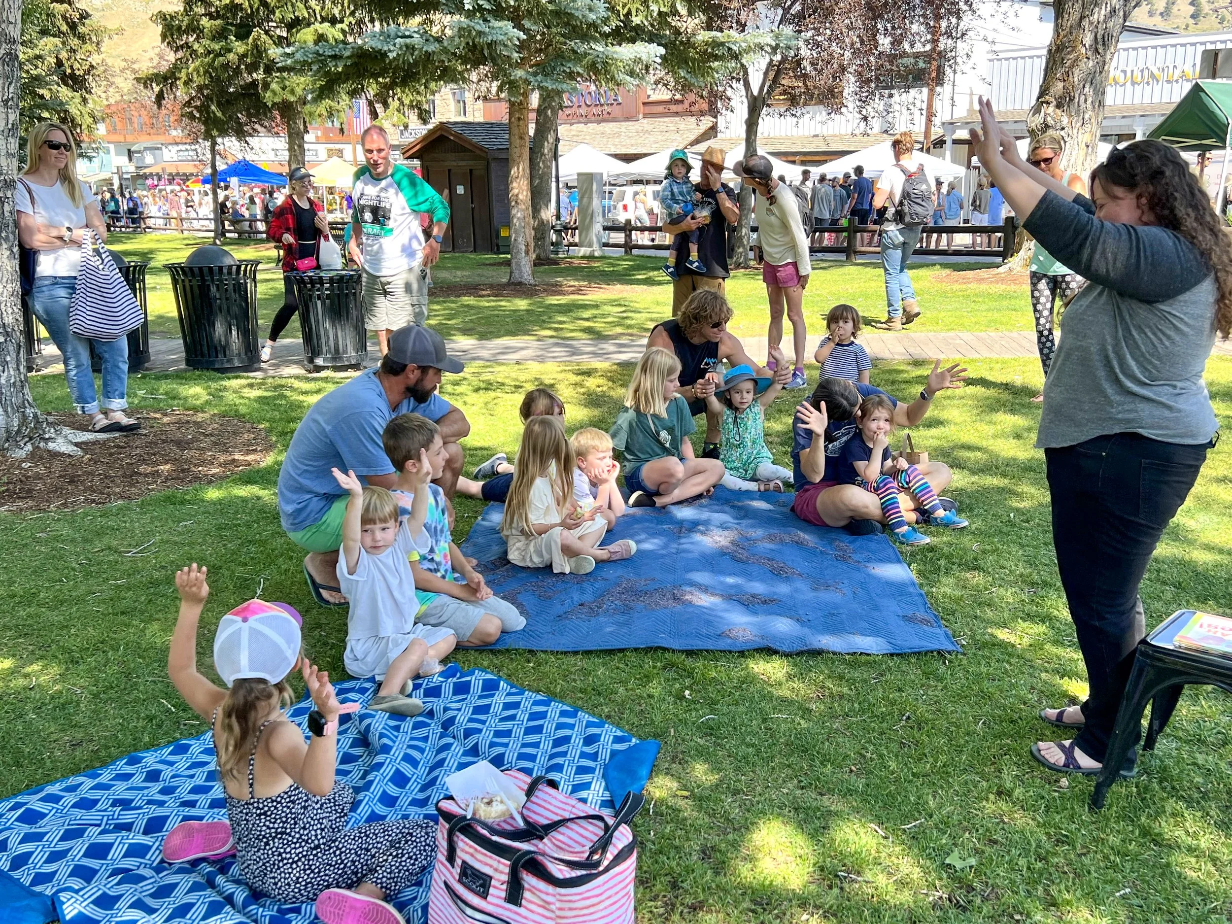 Group of children and adults sitting on blankets in a park, participating in an outdoor storytelling or educational activity, with a woman standing and speaking to the group, and people walking and standing in the background at a fair or community event.