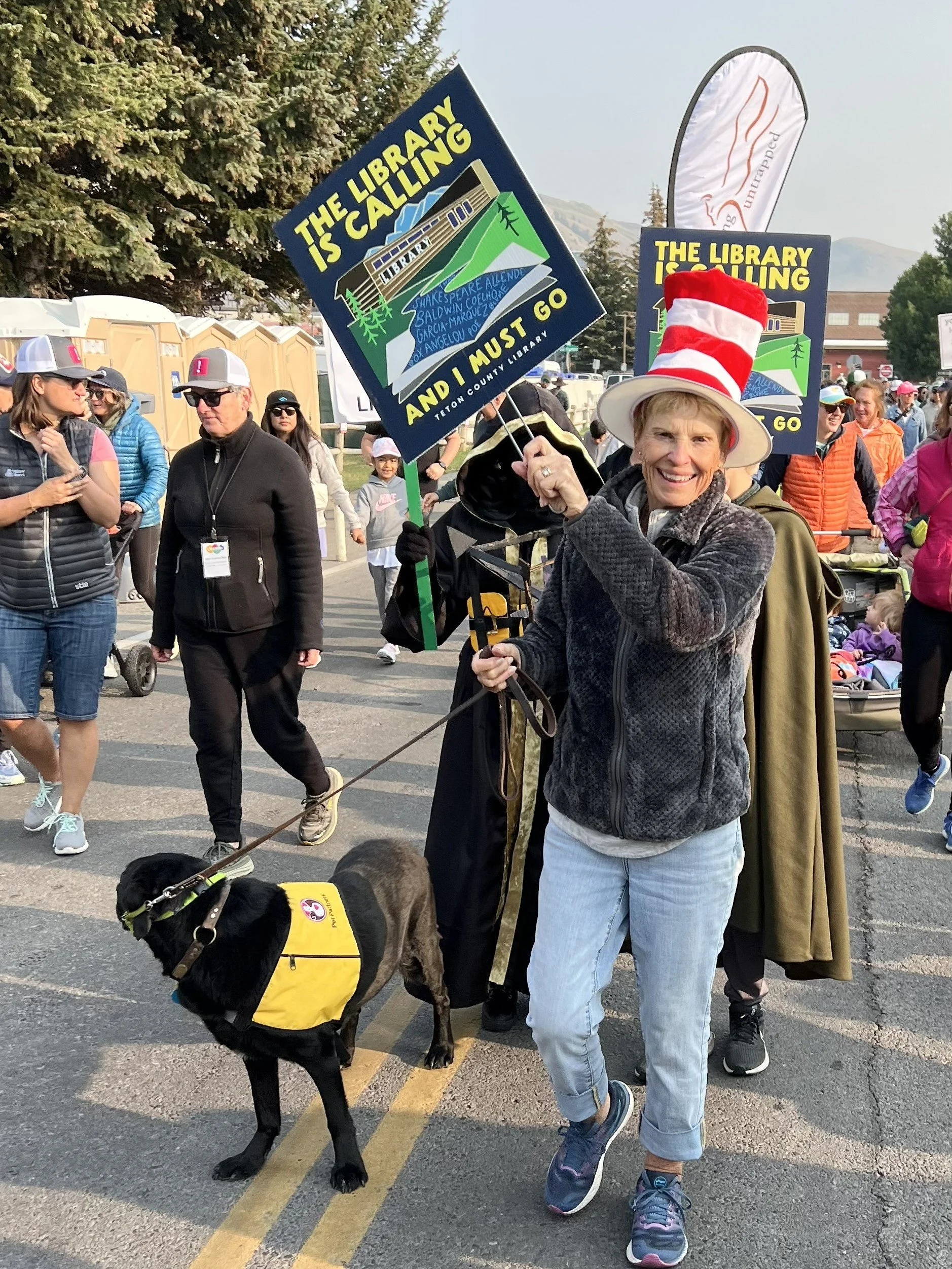 Woman wearing a red and white striped hat holding a sign for the library during a parade. The sign reads: 'The library is calling and I must go.' She is walking a dog in a yellow service vest. Several people are in the background, some wearing hats a