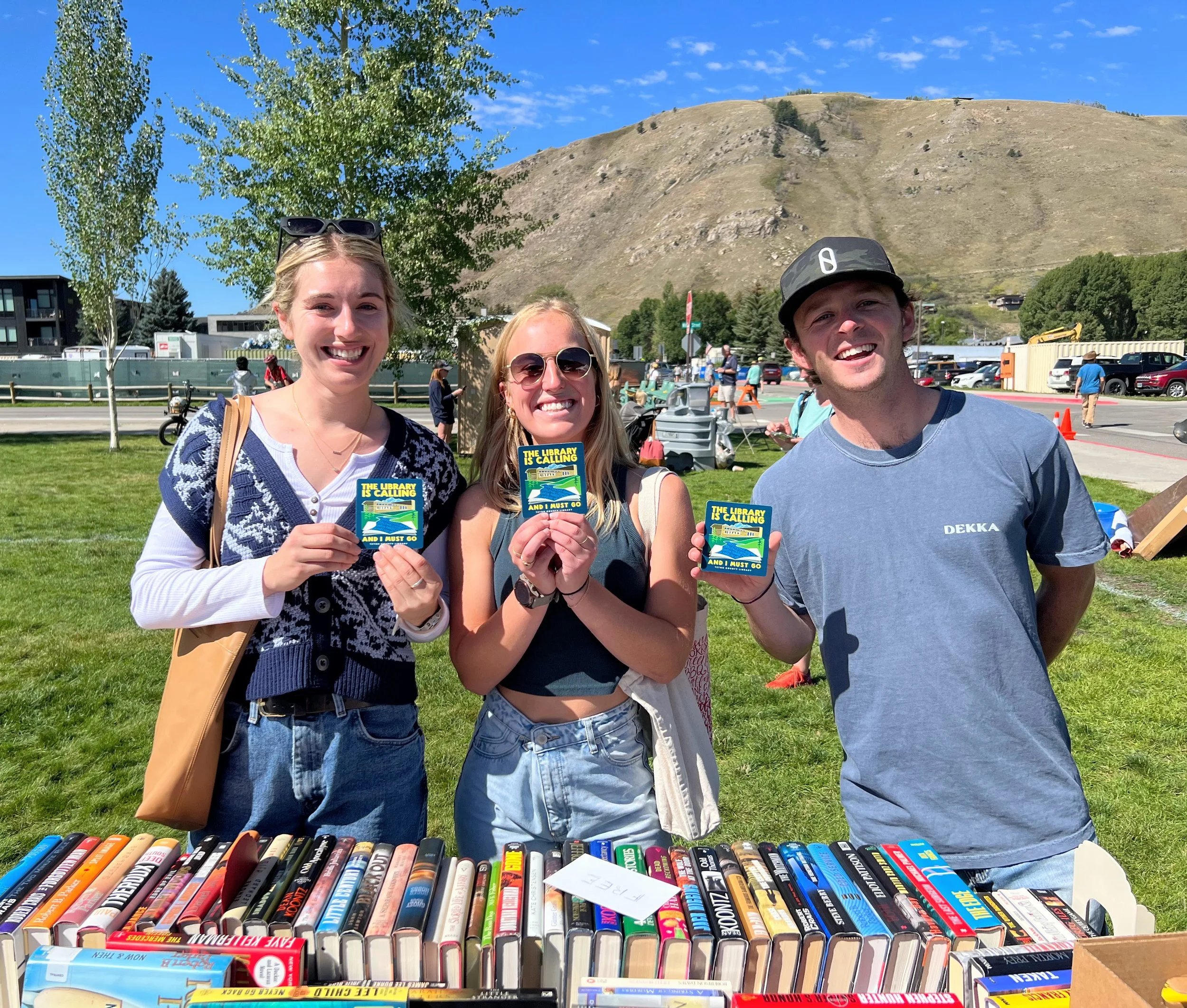 Three smiling young adults holding book sale tags at an outdoor book sale event on a sunny day, with a table of books and a grassy area behind them.
