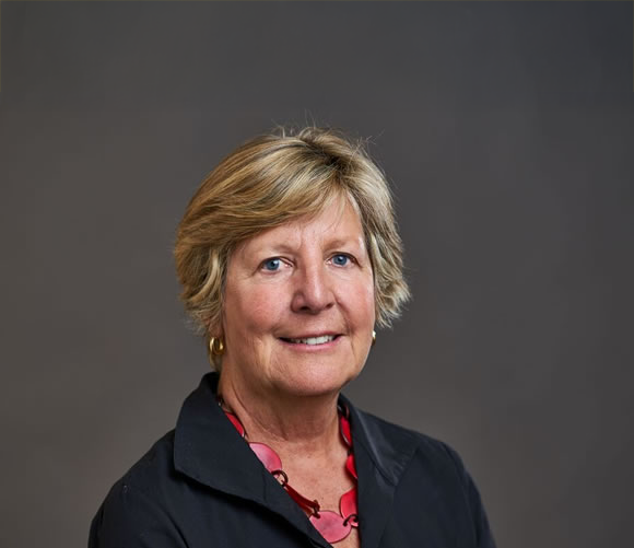 A portrait of a middle-aged woman with short blonde hair, wearing a black jacket and a red necklace, against a neutral gray background.