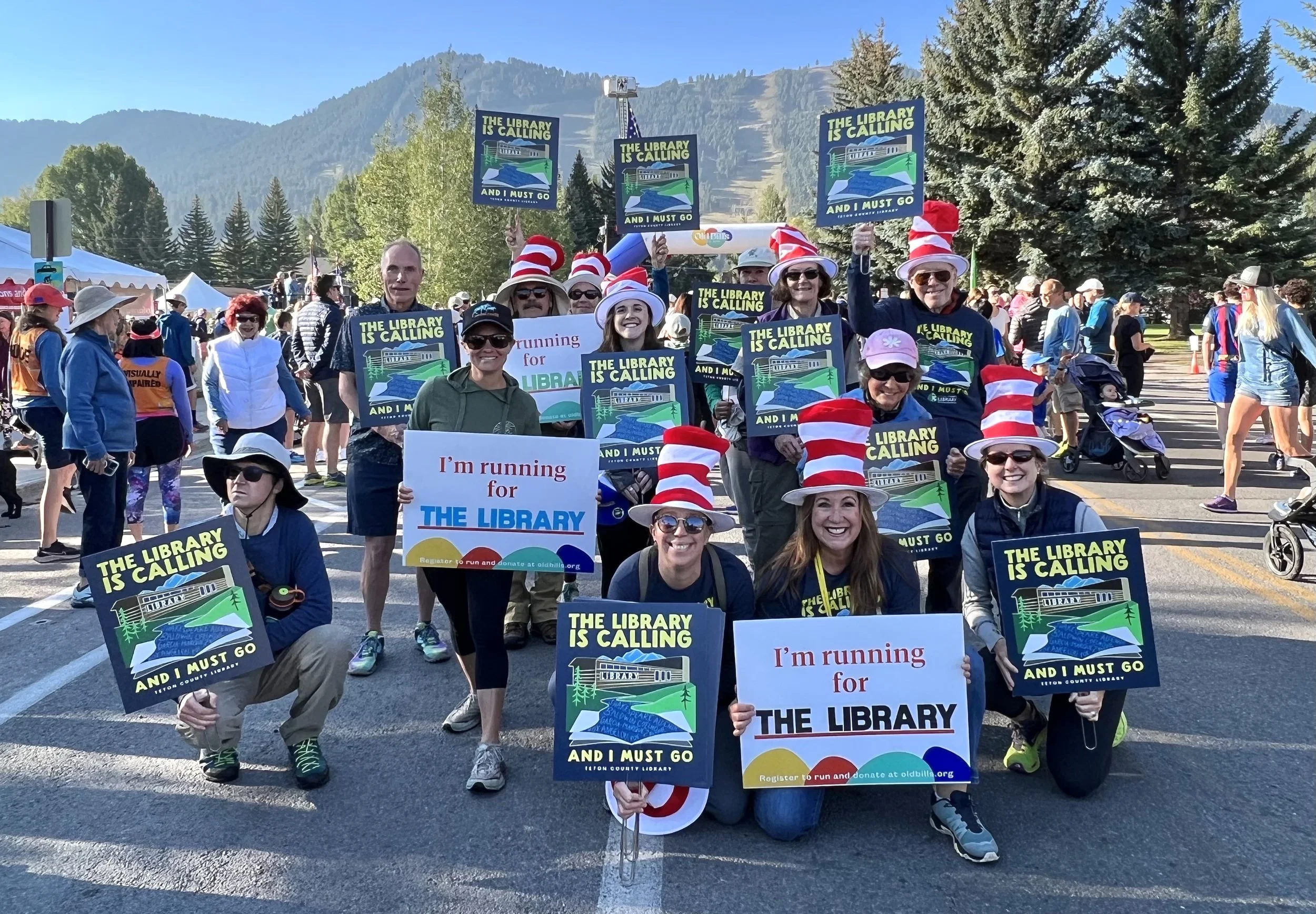 Group of people participating in a parade or rally, holding signs that say 'The library is calling and I must go' and 'I'm running for the library,' wearing Dr. Seuss-inspired hats, sunglasses, and casual clothing, with a mountain landscape and trees in the background.