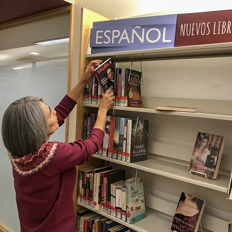 A woman is reaching for a book on a library shelf labeled 'ESPAÑOL' and 'NUEVOS LIBROS', meaning 'Spanish' and 'New Books'. She is middle-aged with gray hair, wearing a maroon sweater. The shelf contains several books, some face out and some spine out, with health and romance genres visible.