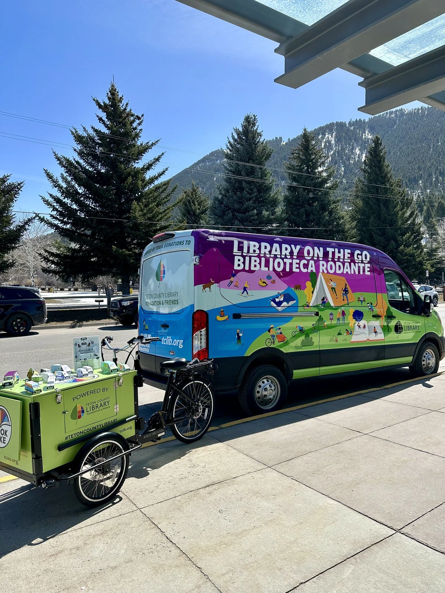 A colorful mobile library van parked outside with the message 'Library on the Go Biblioteca Rodante' on the side, alongside a small bike trailer with the Teton County Library logo, books on display, and a bicycle attached to the trailer. The background includes tall pine trees, mountains, and a clear blue sky.