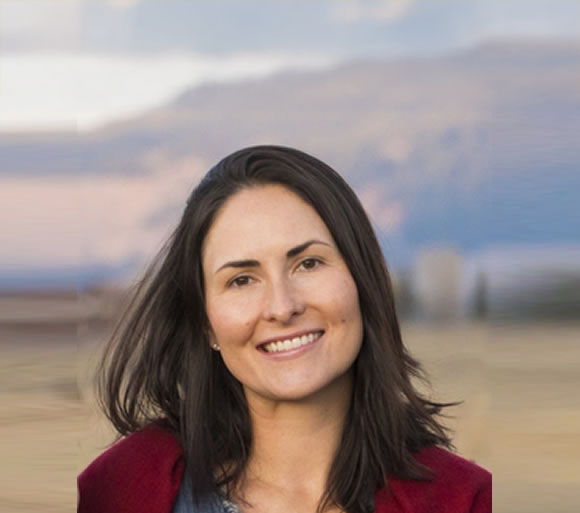 A woman with shoulder-length dark hair smiling outdoors with mountains and a cloudy sky in the background.