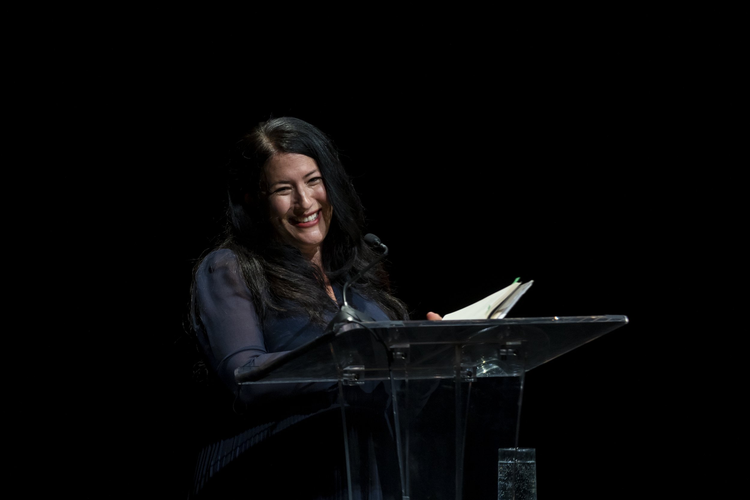 A woman with long black hair and a dark blouse smiling while standing at a transparent podium with a microphone, holding an open notebook, against a black background.