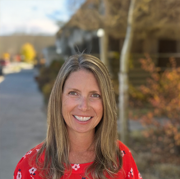 A woman with long blonde hair wearing a red floral dress, smiling outdoors with trees and buildings in the background.