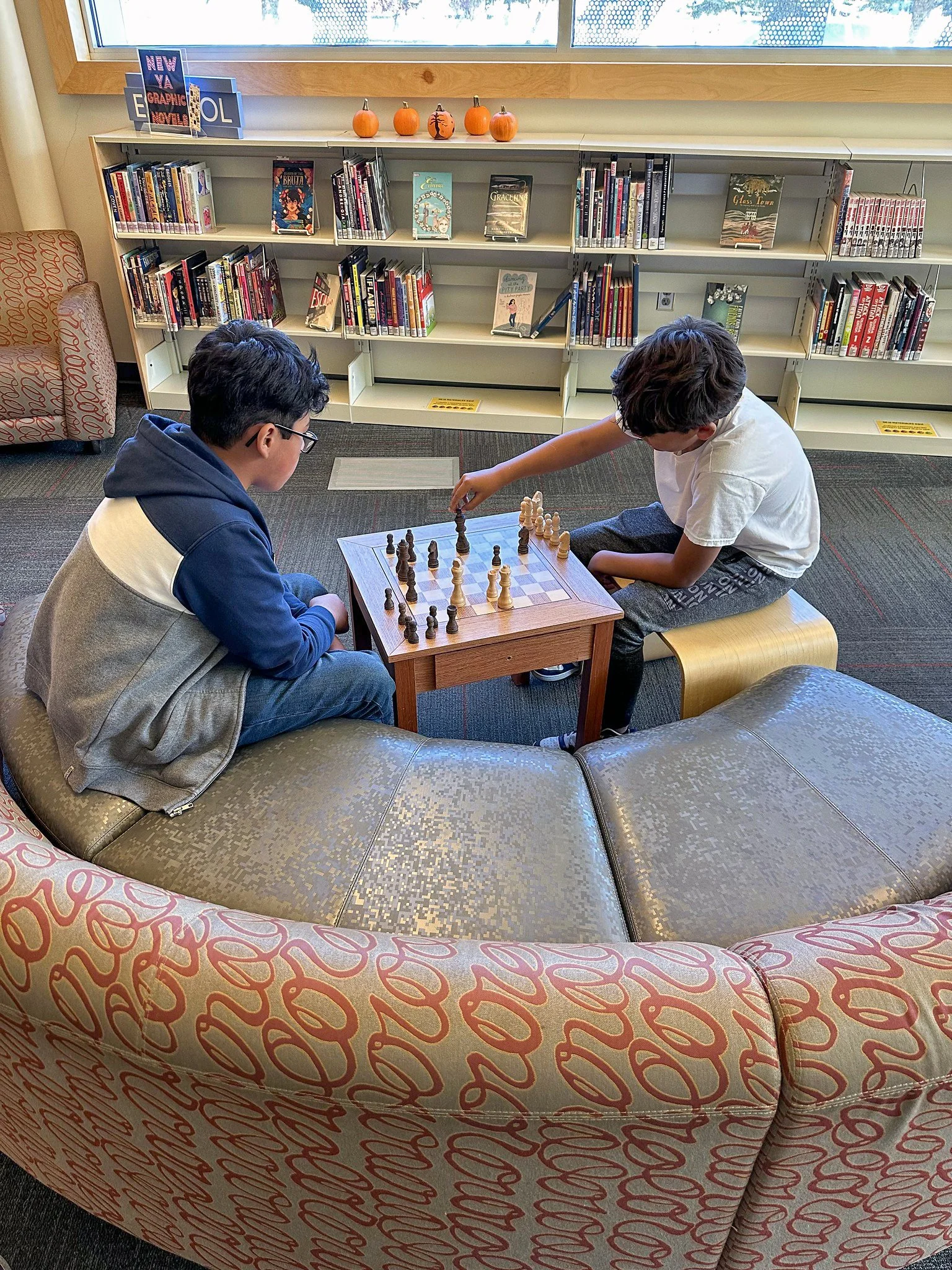 Two children playing chess on a small wooden chessboard in a library, sitting on circular cushioned seats. Bookshelves filled with books are in the background, and a window with pumpkins decorated on the window sill above.