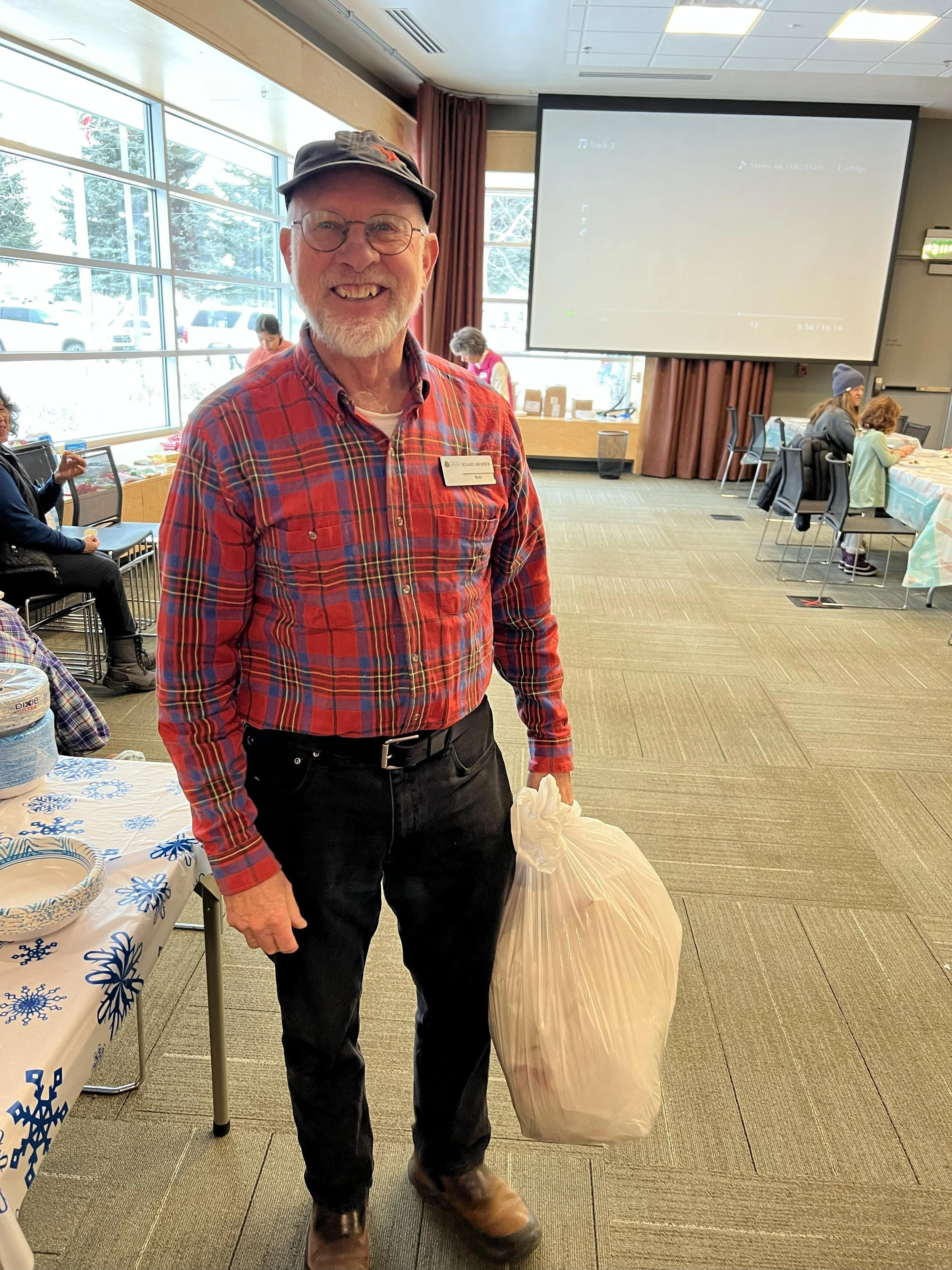 A cheerful elderly man with glasses, a beard, and a black cap, smiling and holding a white plastic bag in a community hall with large windows, tables, and other people in the background.