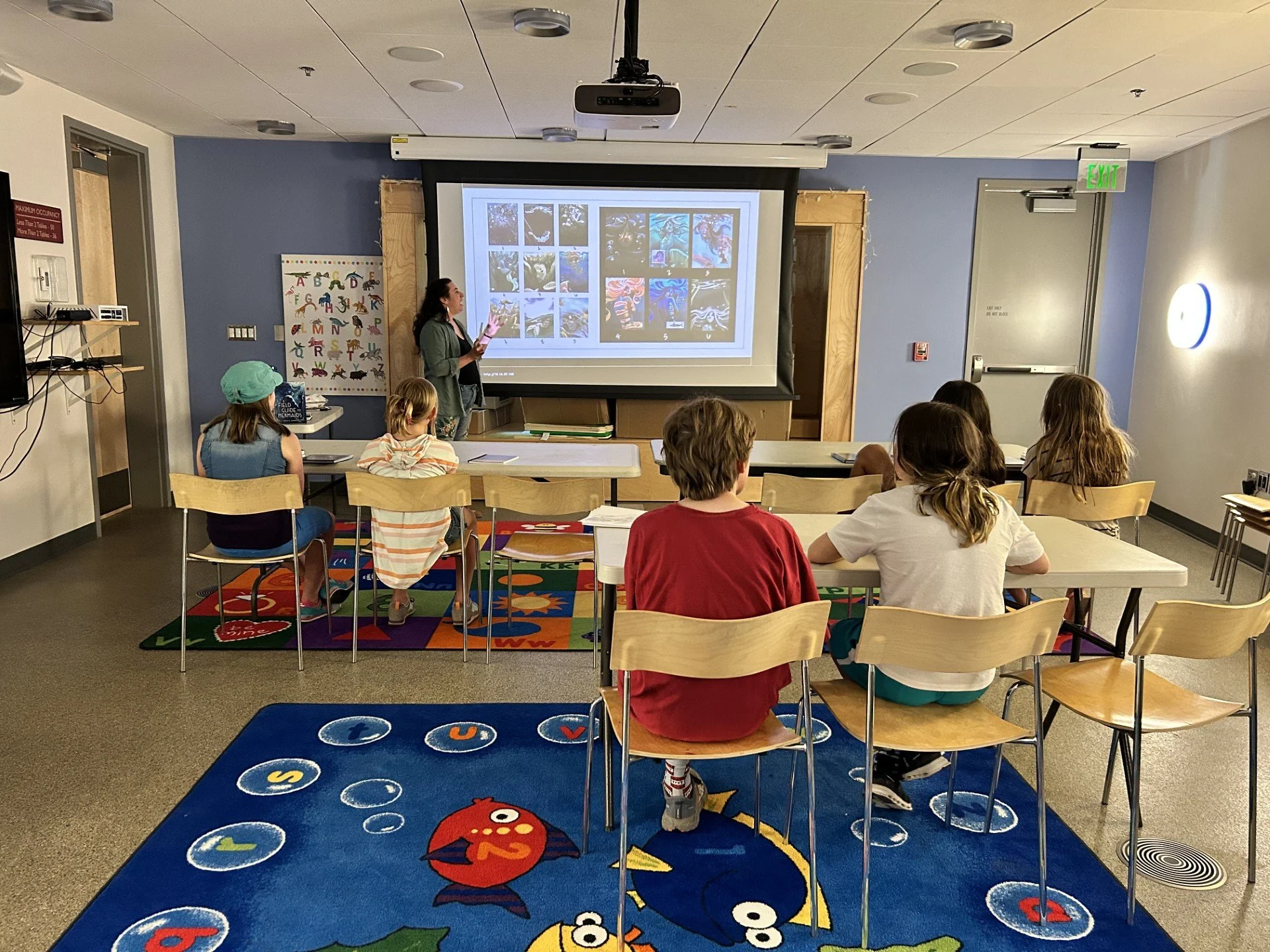 A classroom with children seated at desks watching a teacher present a slideshow on a large screen. The room has colorful carpets, one with fish and another with the alphabet, and a blue wall with a poster of the alphabet in the background.